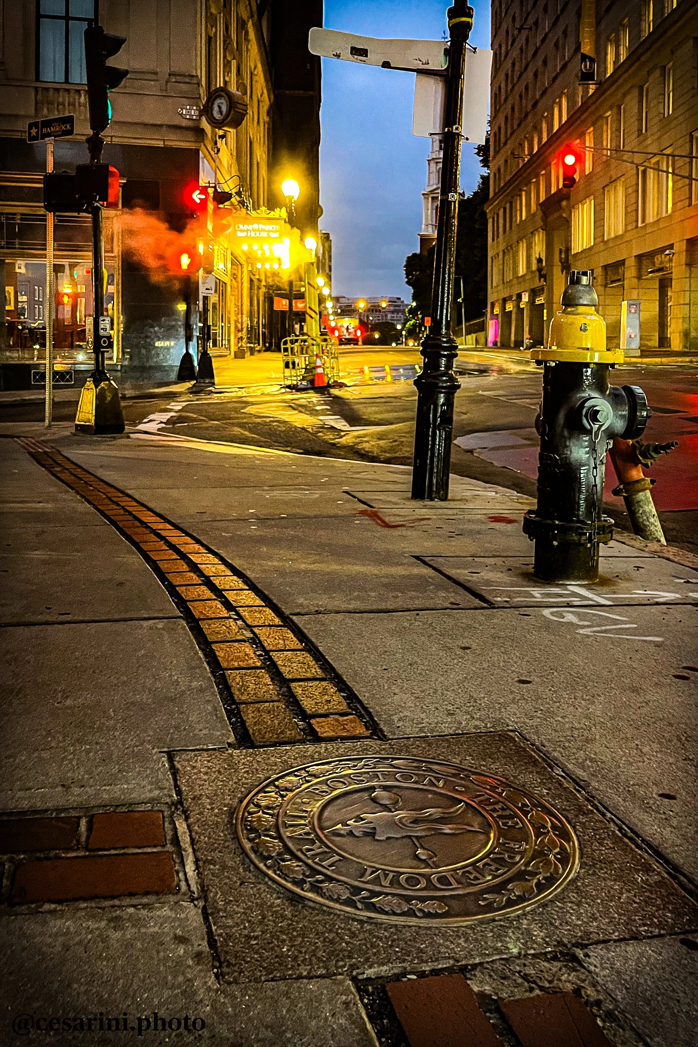 City street at night with traffic lights, steam rising from a vent, and buildings illuminated by streetlights. There are road markings, street signs, and a manhole cover visible.