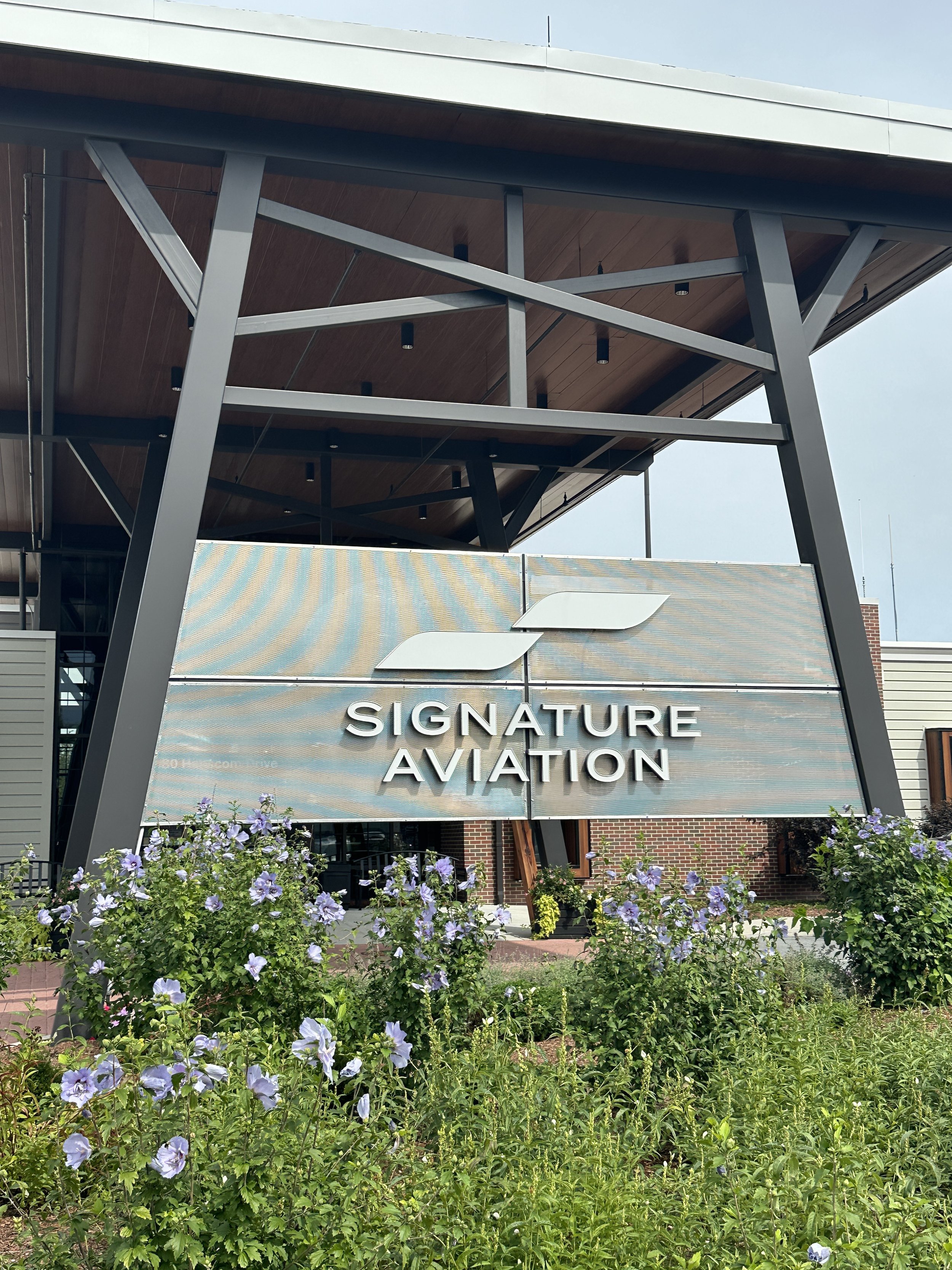 Exterior view of Signature Aviation building with modern architectural design, surrounded by purple flowers and greenery, under a clear sky.