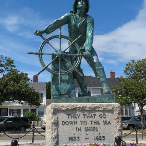 Statue of a woman steering a ship's wheel, with a quote on the base that reads 'They that go down to the sea in ships, 1623-1923,' in a town with houses and trees in the background.