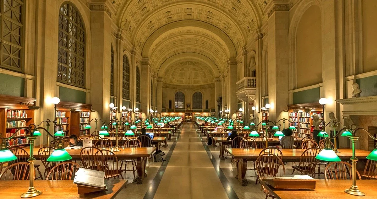A grand library hall with high vaulted ceilings, tall windows, and rows of wooden tables with green-shaded lamps. People are reading and working at the tables.