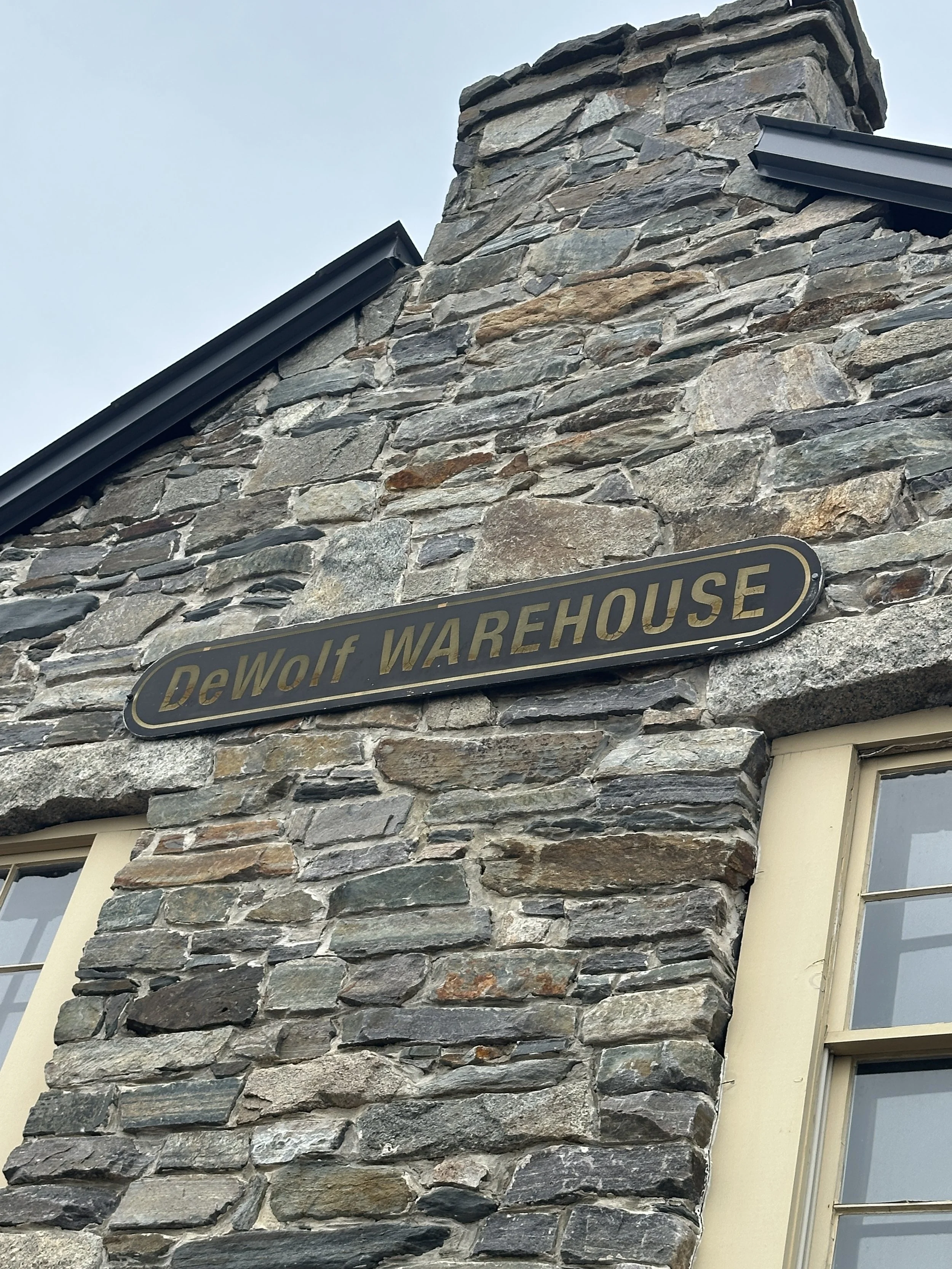 Close-up of a stone chimney on a house with a metal sign that reads 'DeWolf WAREHOUSE' mounted on the wall below the chimney.