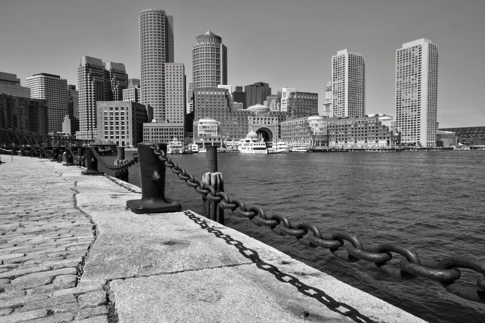 Black and white photo of a city skyline across a harbor, with tall skyscrapers and boats docked along the waterfront,. A stone pier with a chain barrier is in the foreground.