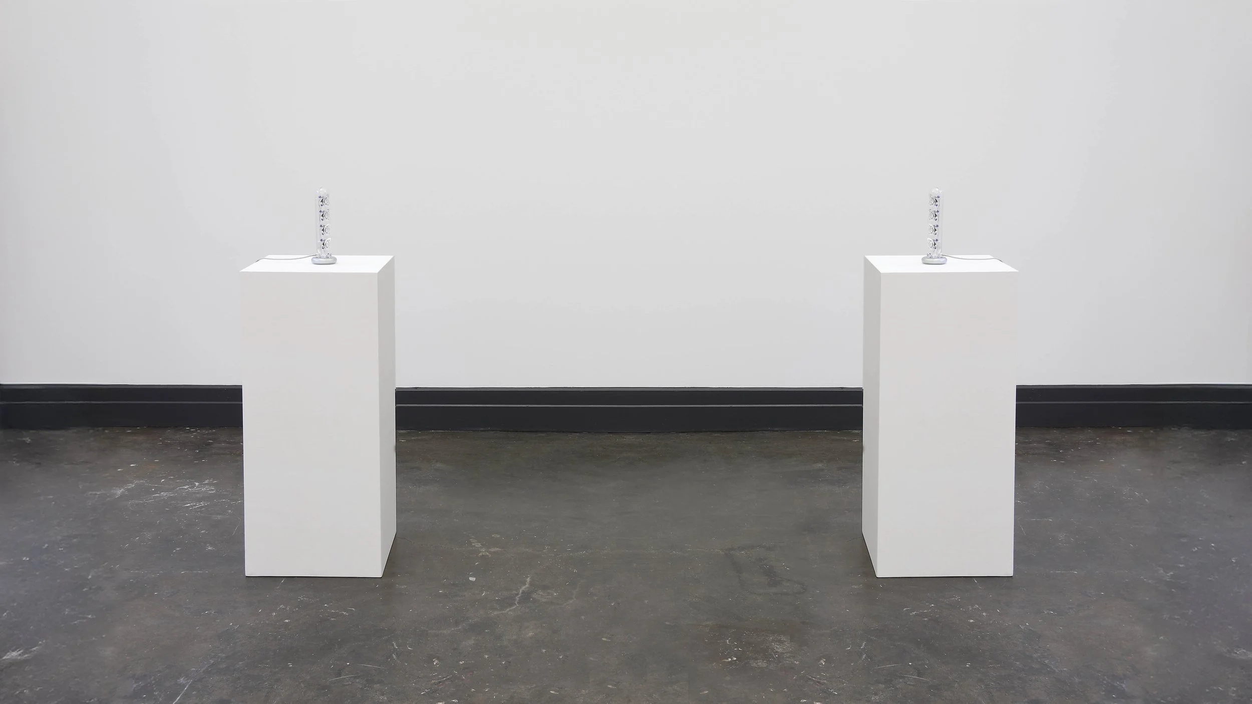 Two white pedestals face each other with a small speaker on top, arranged in a gallery or exhibition space with a plain white wall and a black baseboard.