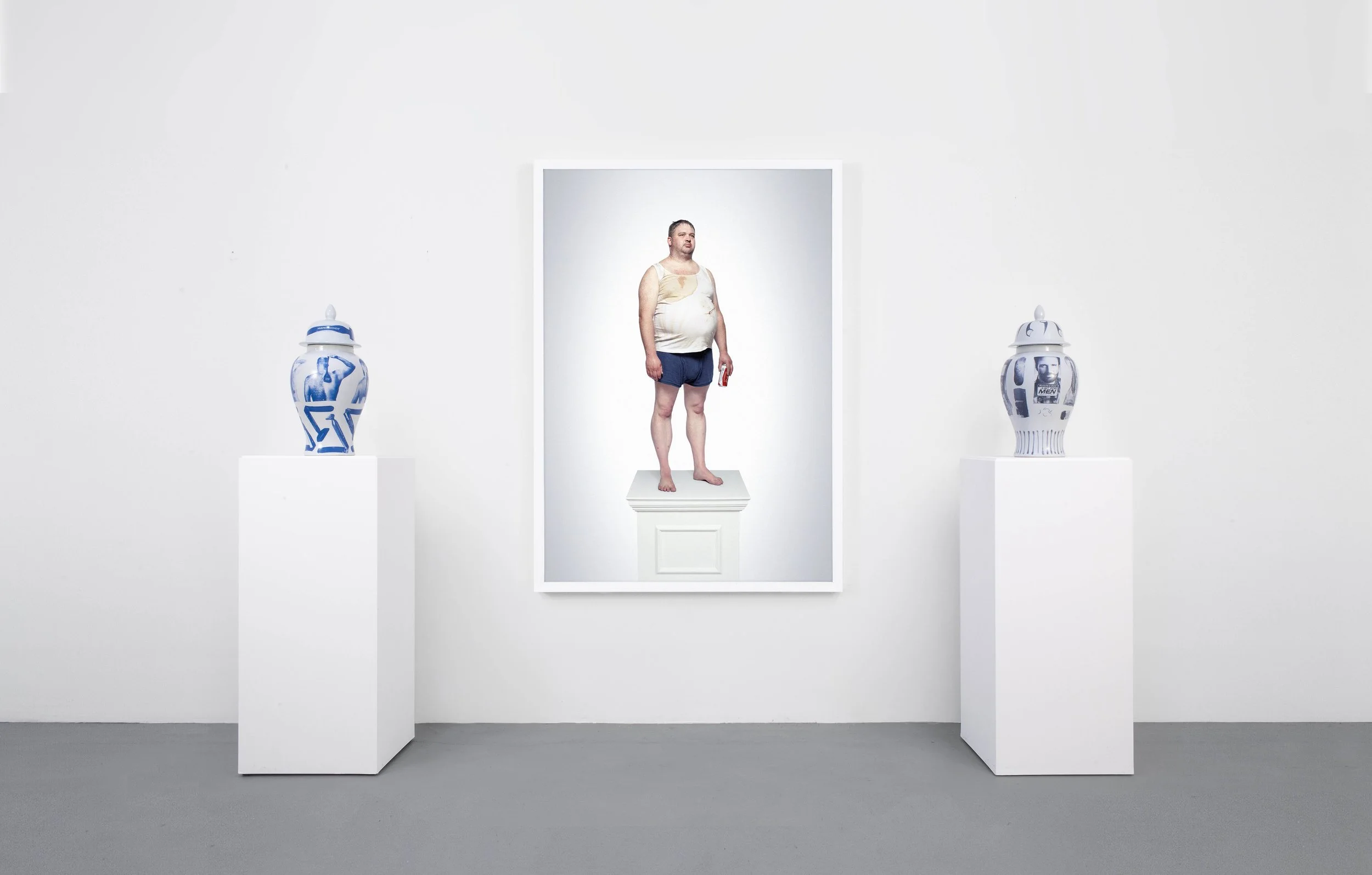 An art installation featuring a photographic portrait of a slob standing on a pedestal, flanked by two decorative vases on white pedestals, in a minimalist gallery setting.
