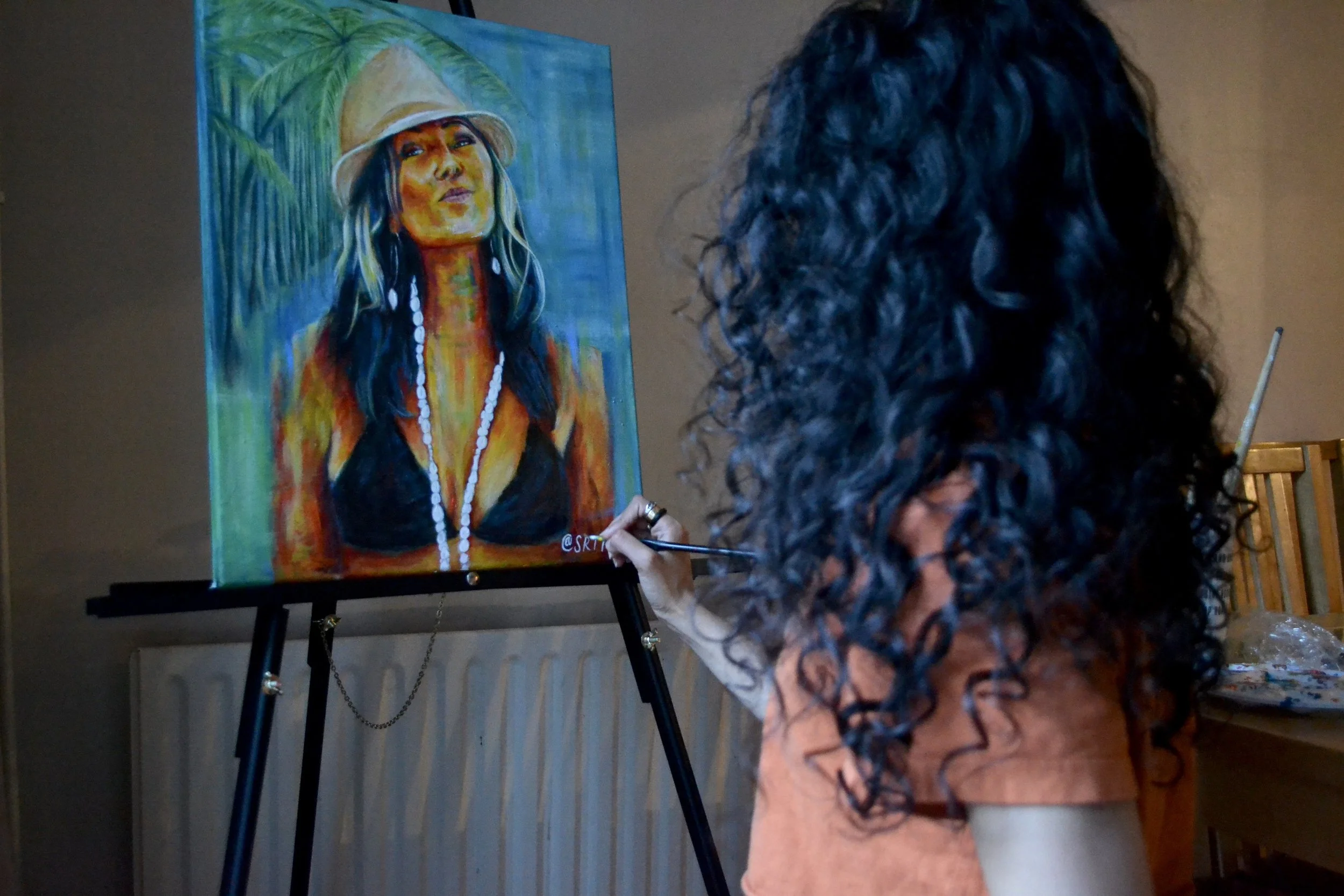 Artist with curly dark hair painting a portrait of a woman Anuhea wearing a hat, black top, and shell necklace on an easel.