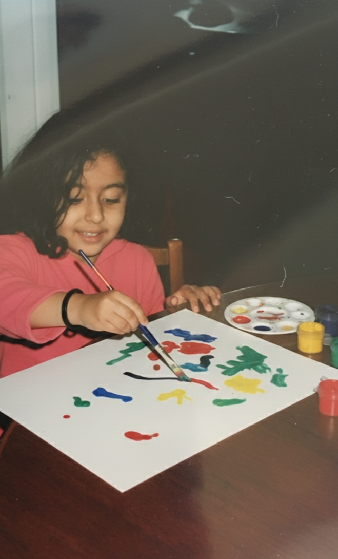 A young girl with curly hair wearing a pink shirt is painting with bright colors on a white sheet of paper. She is using a paintbrush and has a palette of paint nearby.