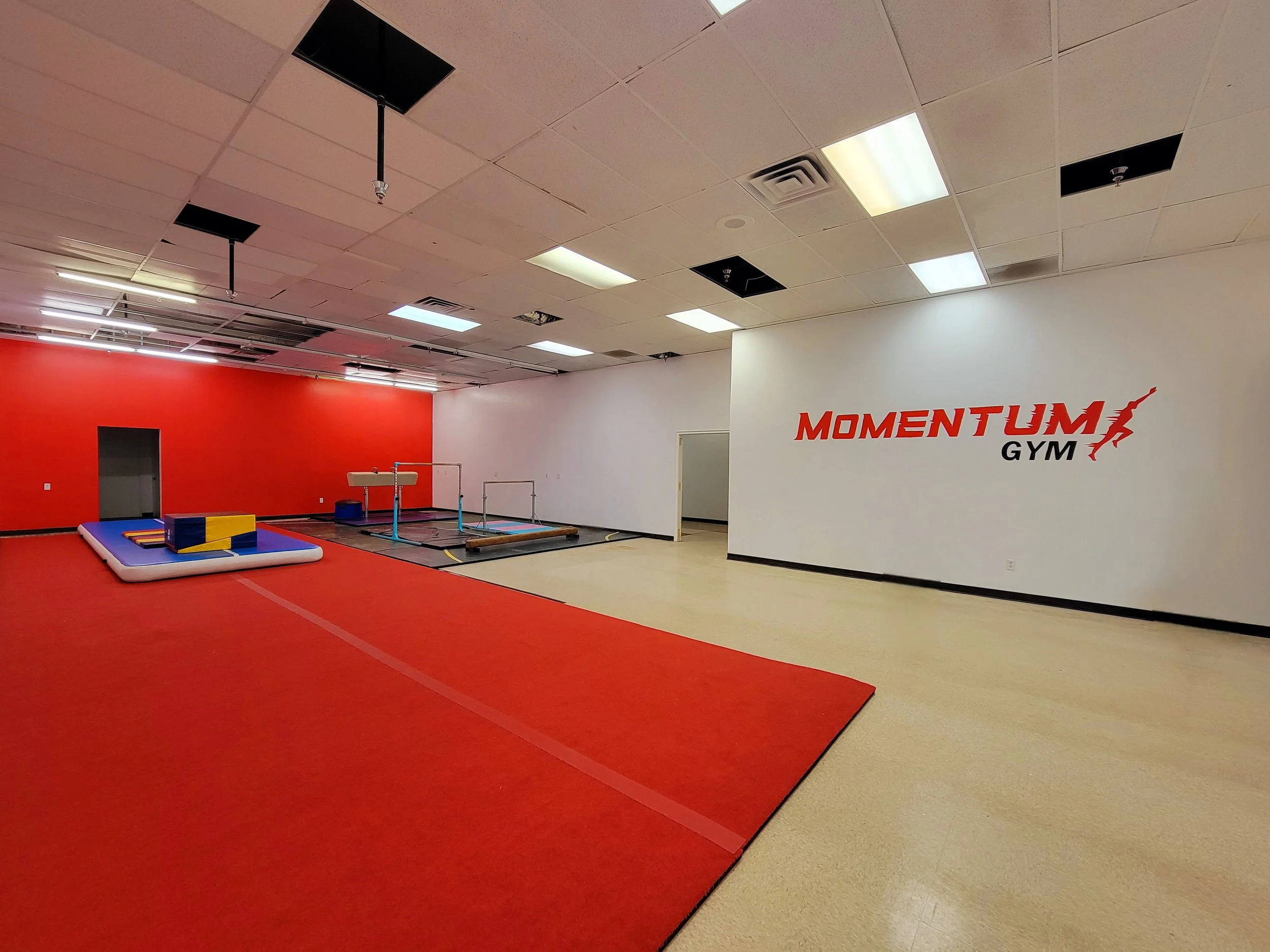 Interior of a gym named Momentum Gym with red and beige flooring, white and red walls, and workout equipment including mats and balance beams.