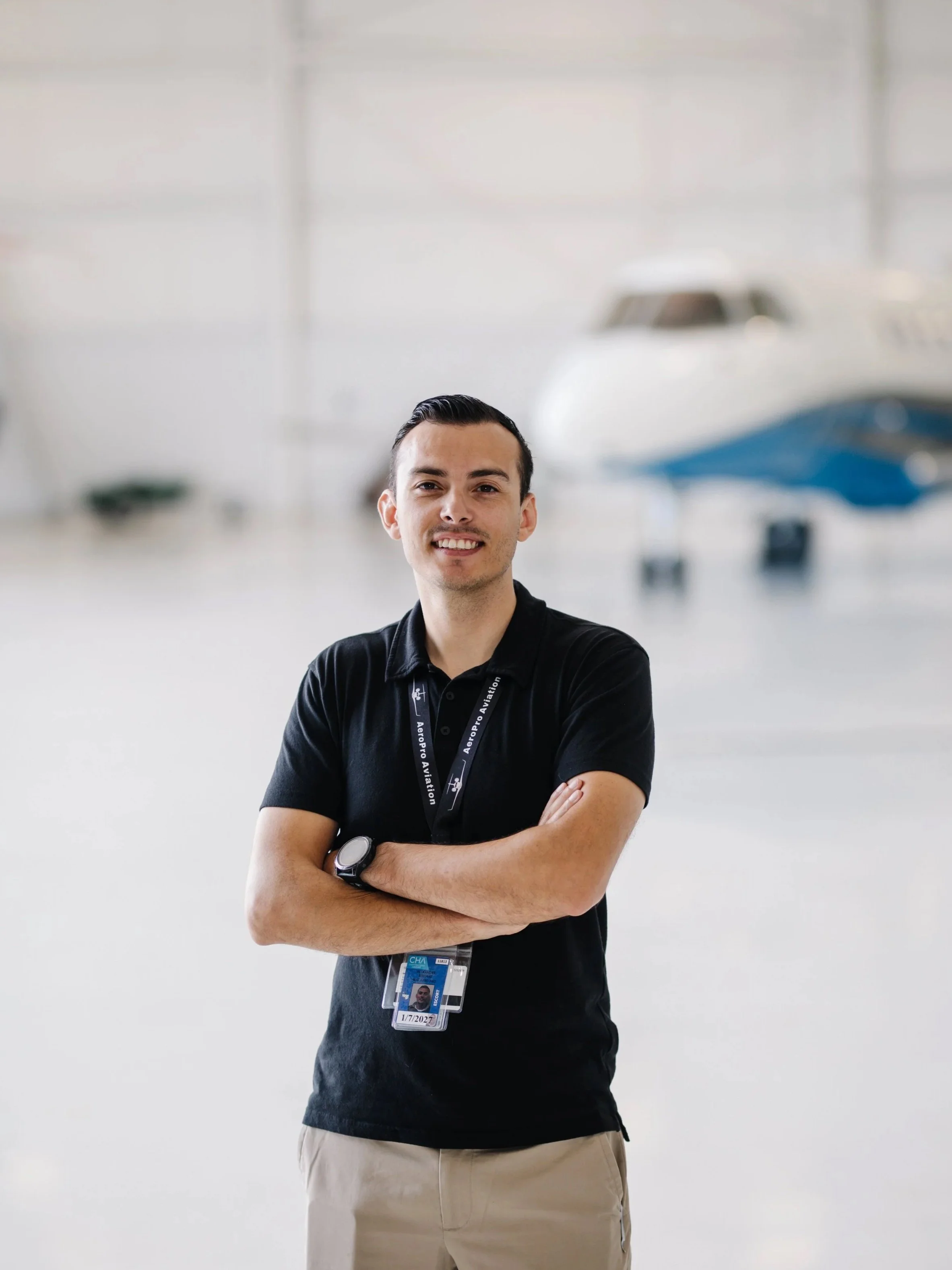 A young man wearing a black polo shirt with a badge around his neck stands with arms crossed in front of a blurred background of an aircraft hangar with airplanes.