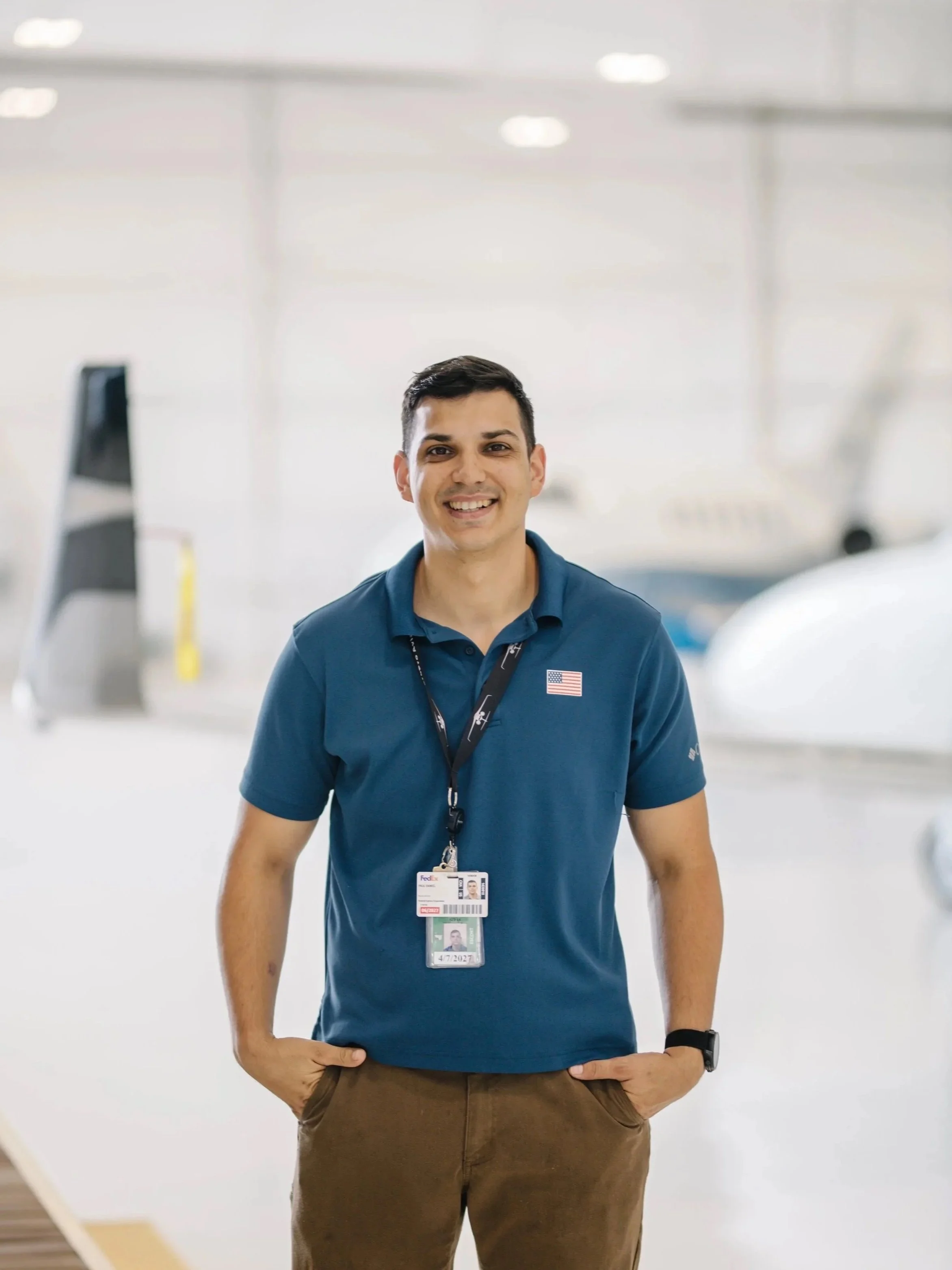 A smiling man wearing a navy blue polo shirt with an American flag patch, brown pants, a badge on a lanyard, and a black wristwatch, standing in an airport hangar with airplanes in the background.