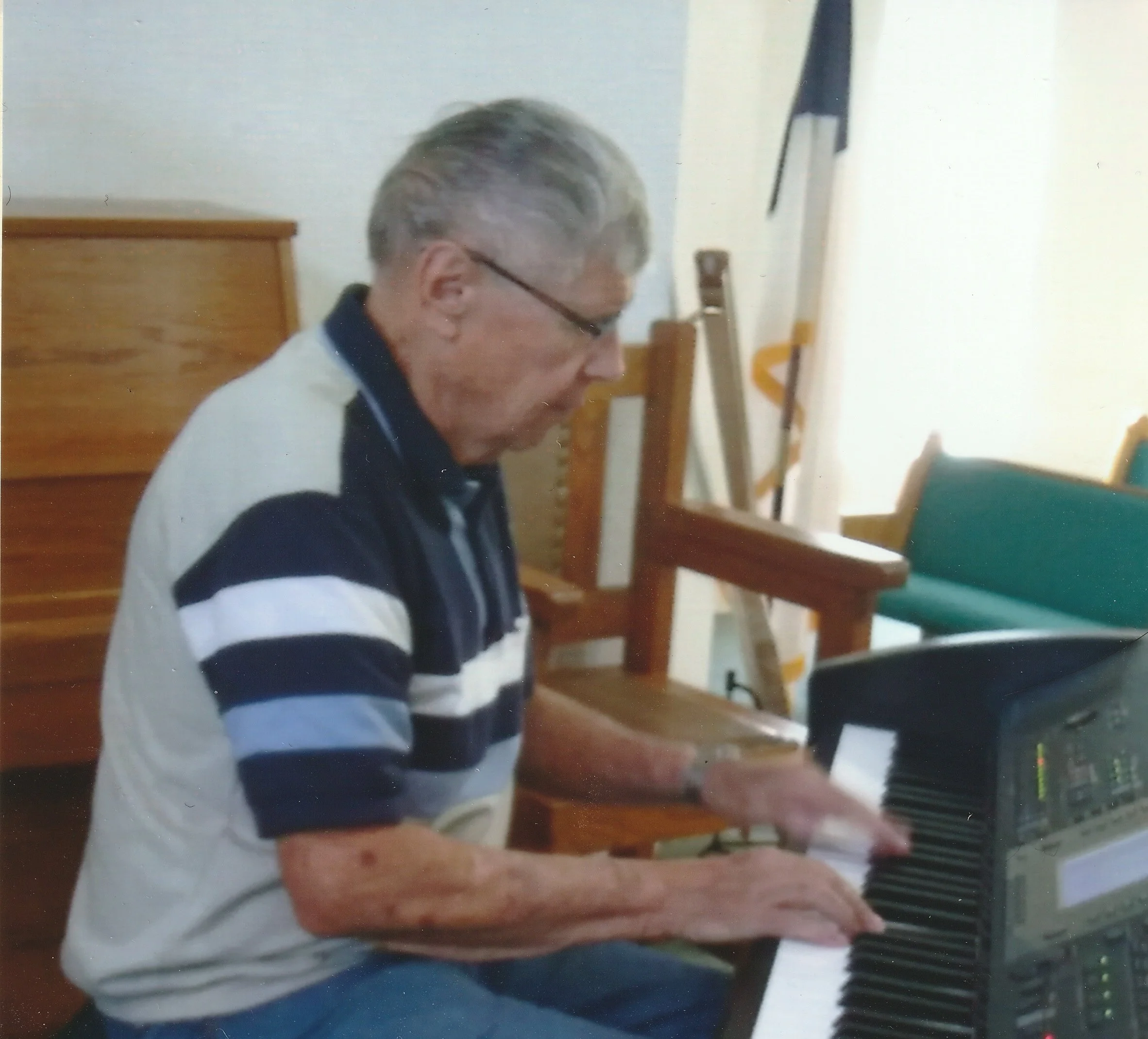 An elderly man with gray hair and glasses playing an electronic keyboard in a room.