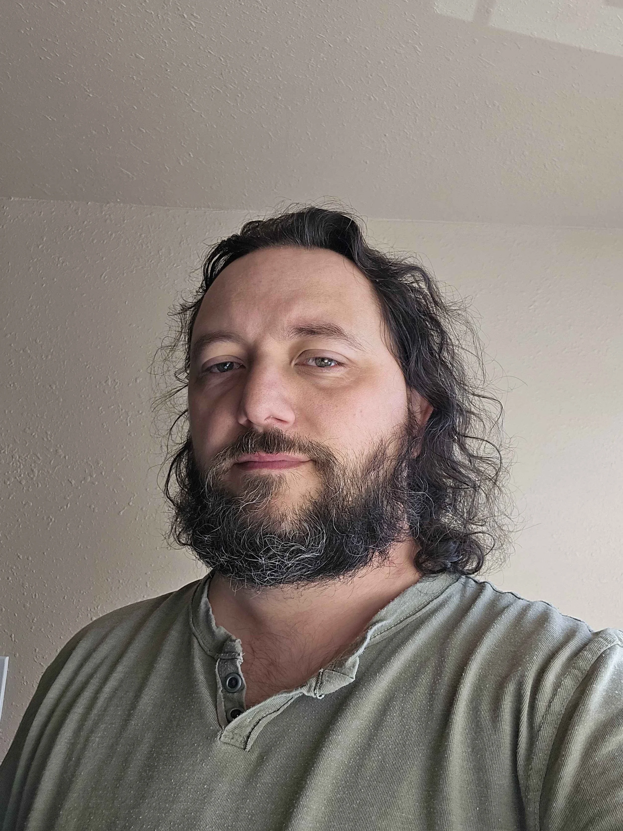 A man with long dark hair and a full beard taking a selfie indoors against a beige textured wall.