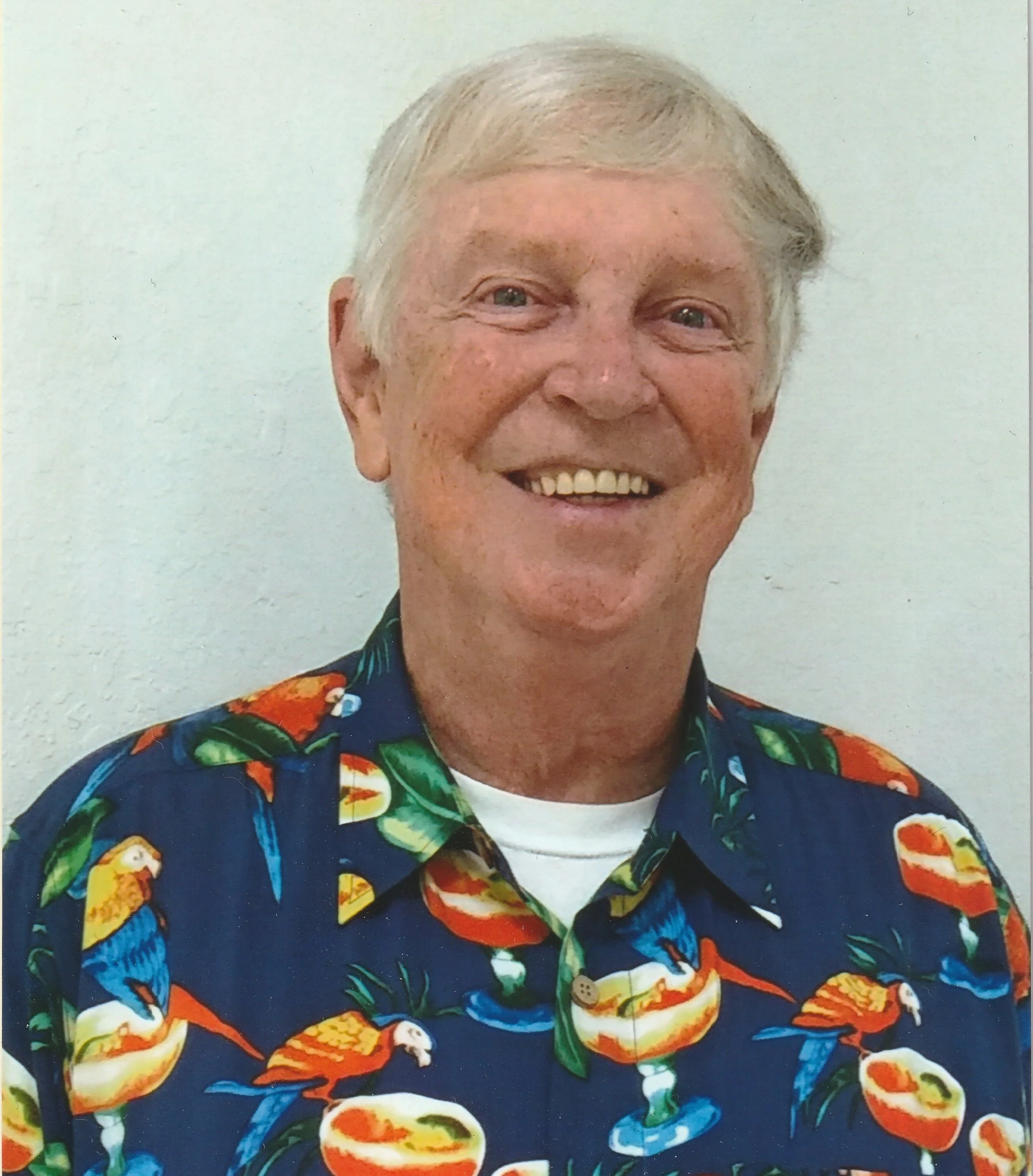 A smiling older man with light hair wearing a colorful Hawaiian shirt with parrots and fruit.