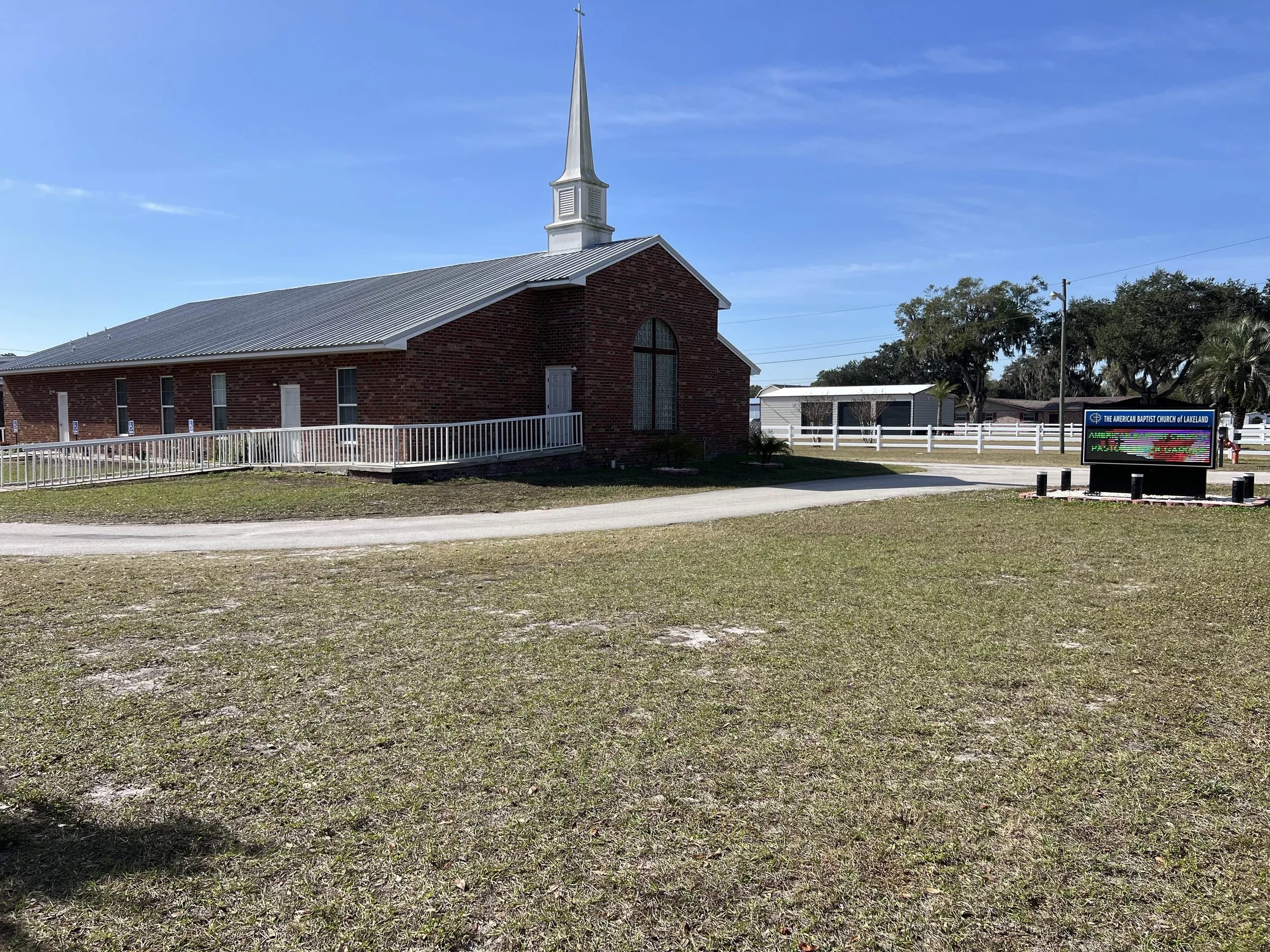 A red brick church with a white steeple on a bright, sunny day. The church has a white railing along the front and side. In front, there is an electronic signboard with a blue header. It's the American Baptist Church of Lakeland, FL.