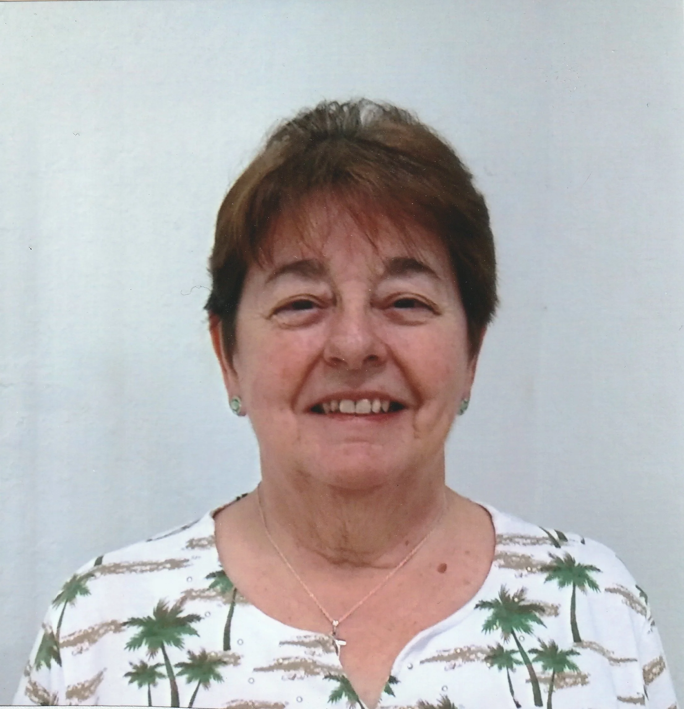 Smiling woman with short brown hair, wearing a white shirt with palm tree prints, earrings, and a necklace.