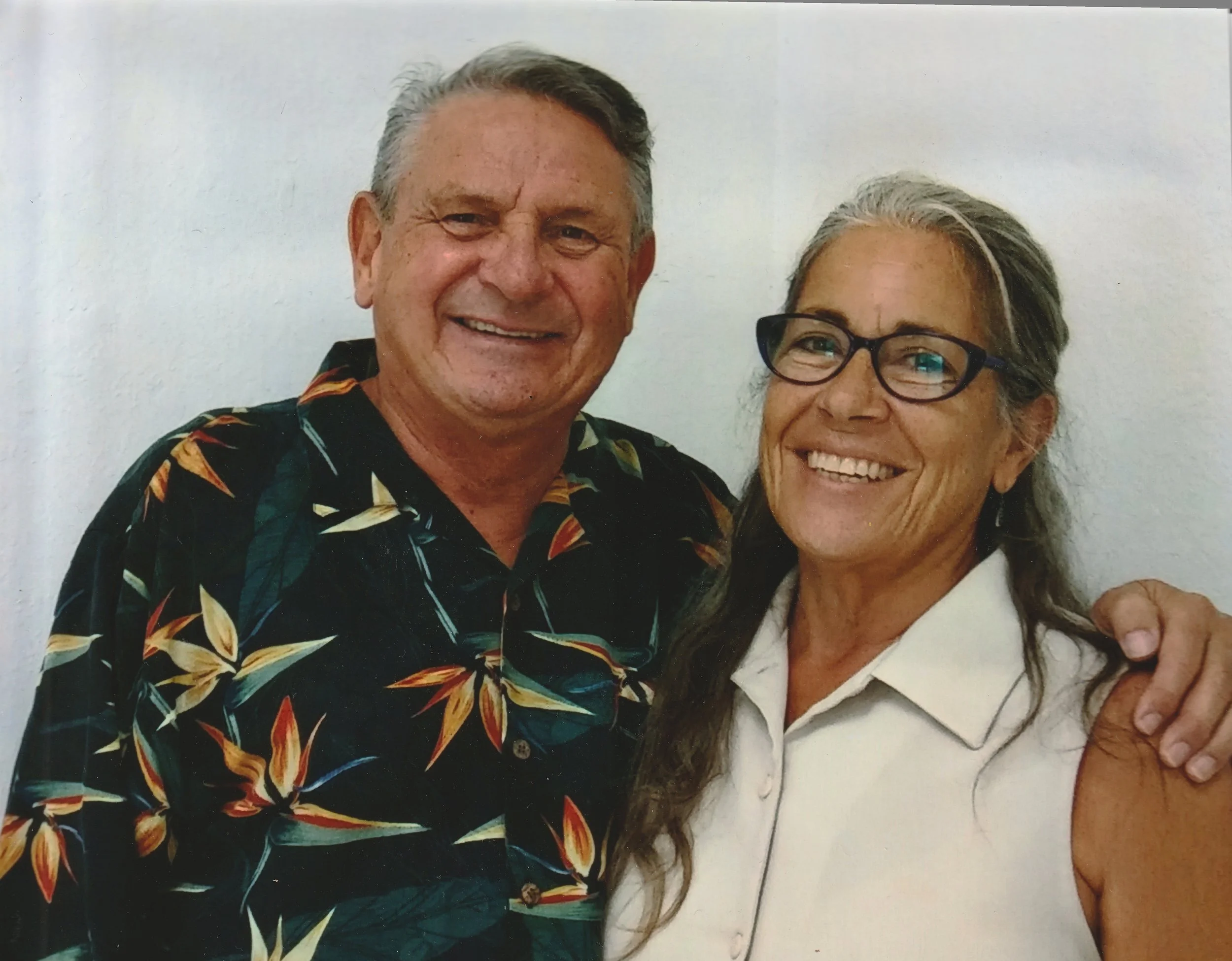 A man and a woman smiling and posing together, with the man wearing a dark floral Hawaiian shirt and the woman wearing a white sleeveless blouse and glasses.
