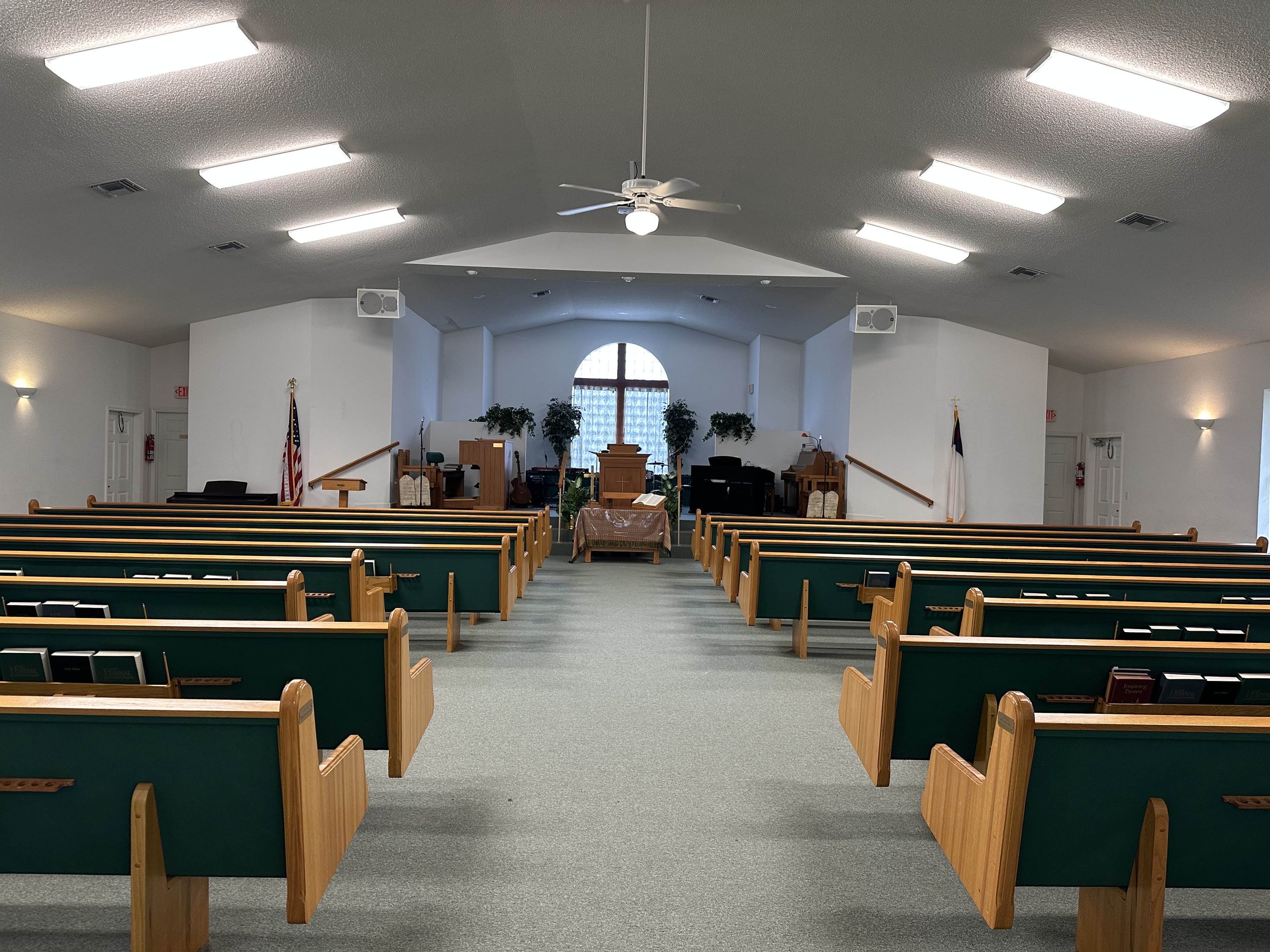 Interior of a church sanctuary with wooden pews facing the altar, a large window behind the altar, potted plants, and church furniture, under ceiling lights.