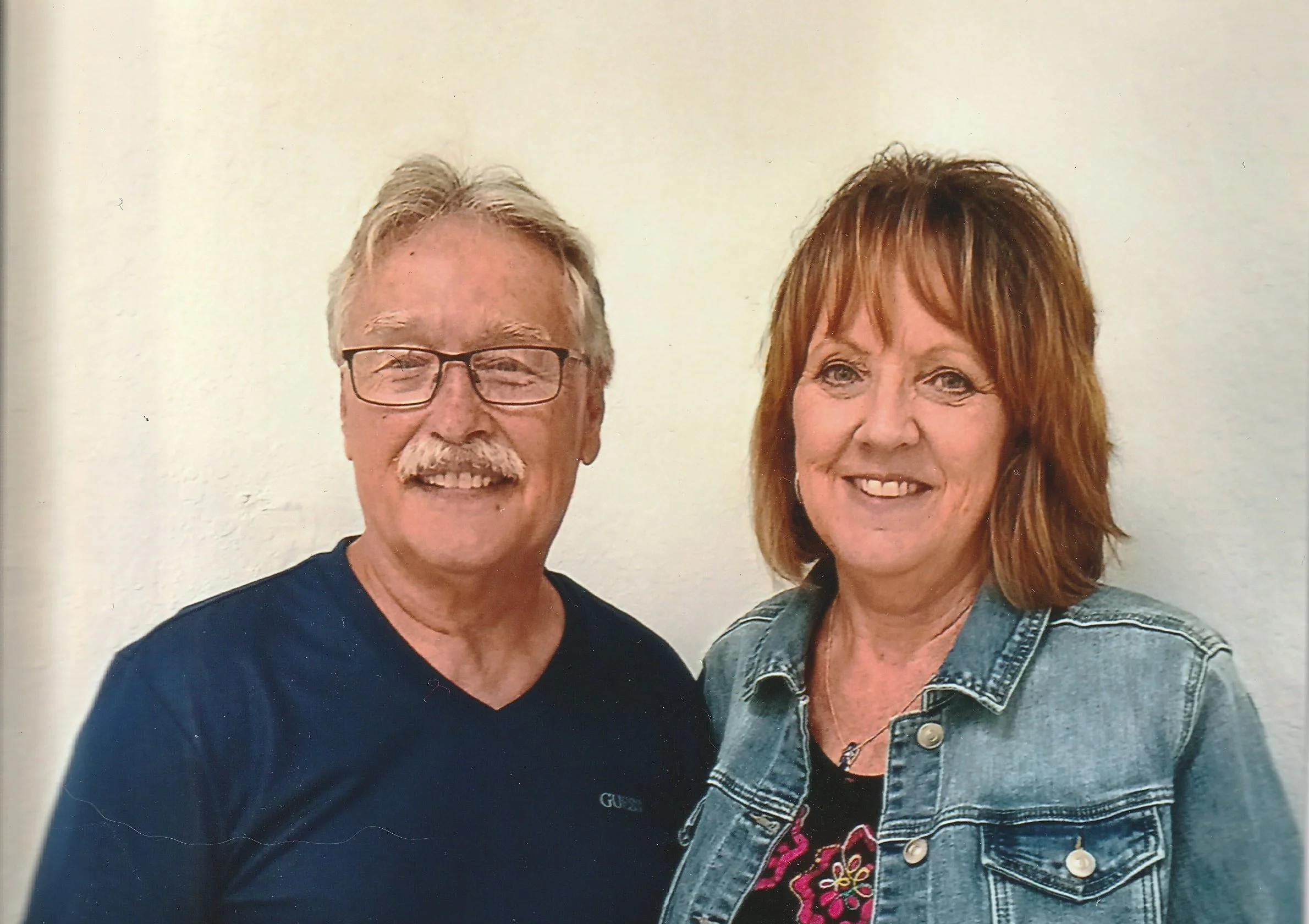 An older man with gray hair, glasses, and a mustache wearing a dark shirt, and an older woman with shoulder-length red hair wearing a denim jacket and a floral shirt, both smiling and standing against a plain light-colored wall.