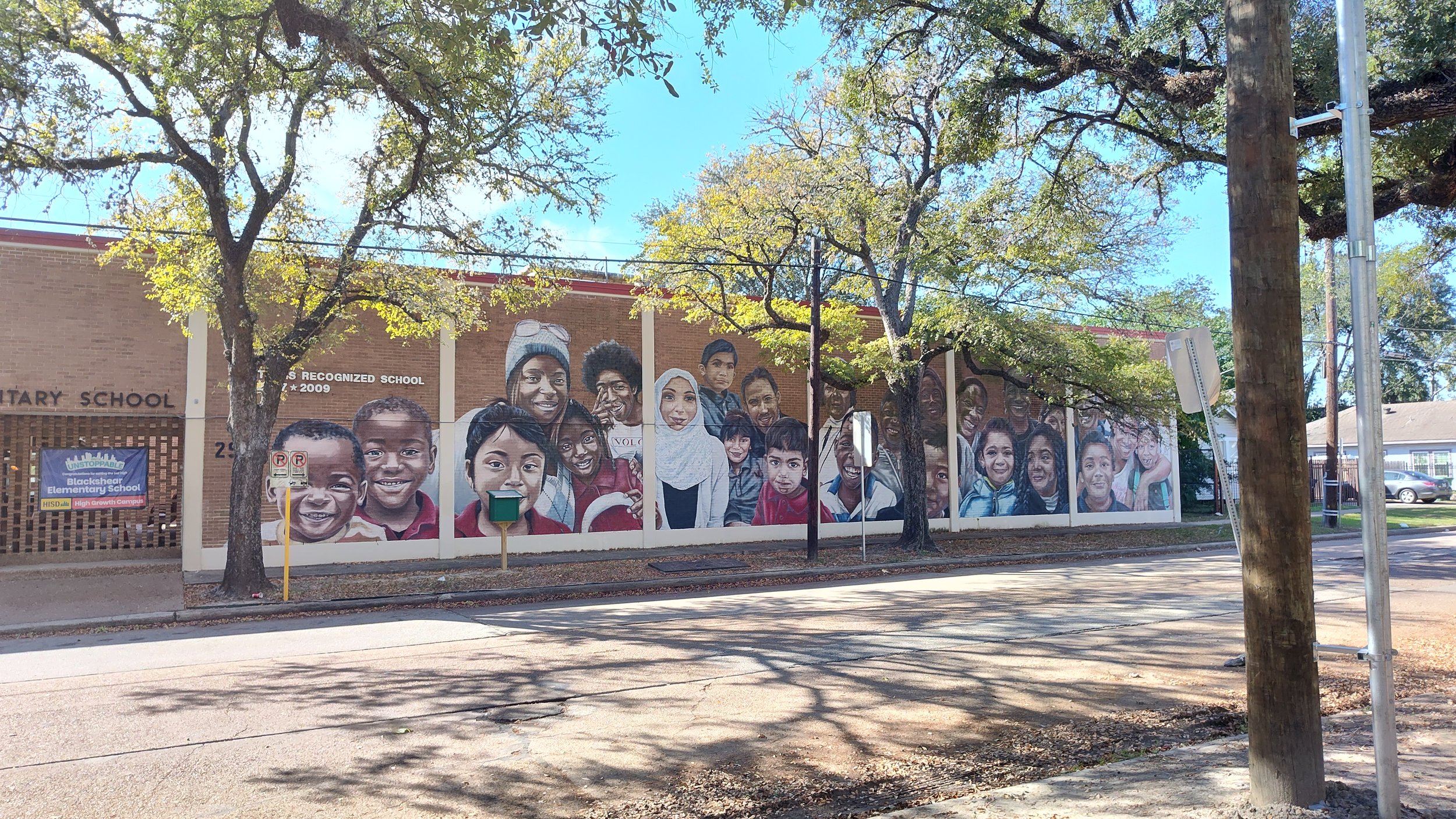 A mural of diverse children and adults painted on the side of a brick school building, with trees and a street in the foreground.