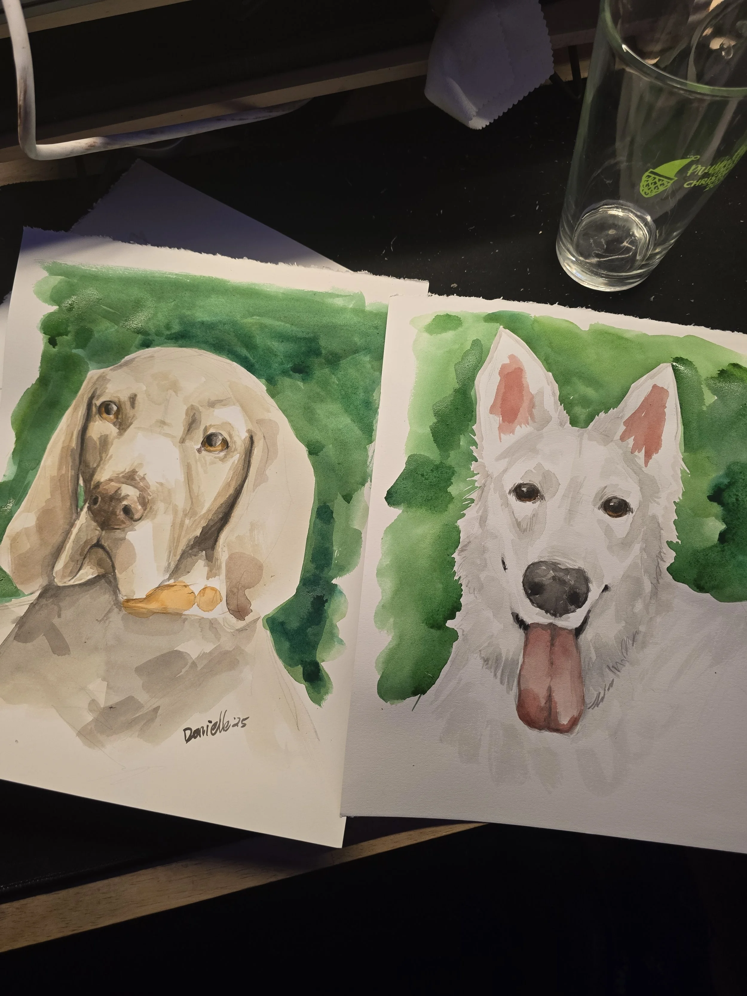 Watercolor paintings of a Labrador retriever and a Siberian husky on a table with a glass and a napkin nearby.