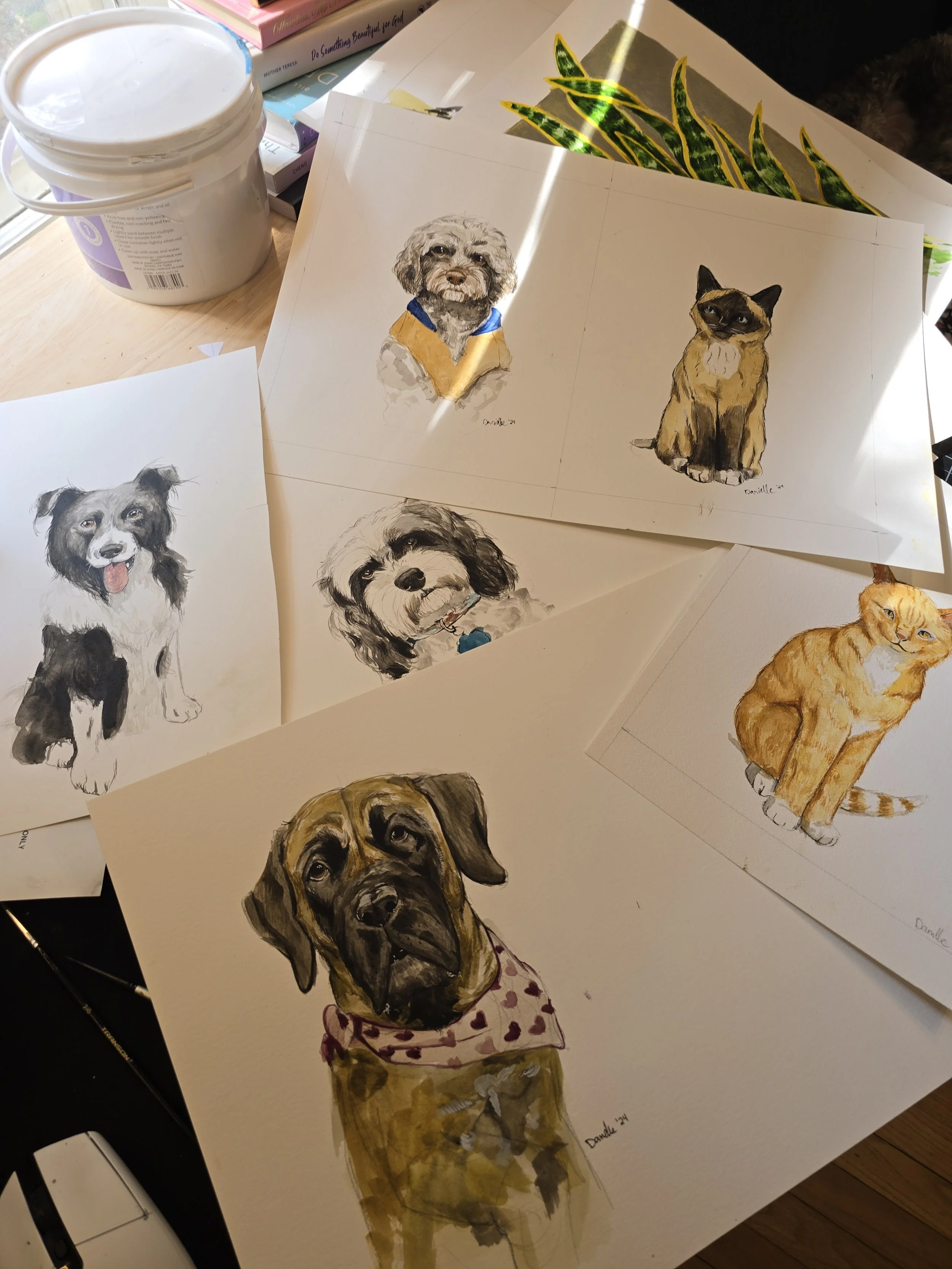 Collection of watercolor dog and cat portraits on paper, placed on a desk with art supplies and books. The portraits include a fluffy dog with a yellow and blue shirt, a black and brown dog with a pink bandana, a cat with a serious expression, a smal