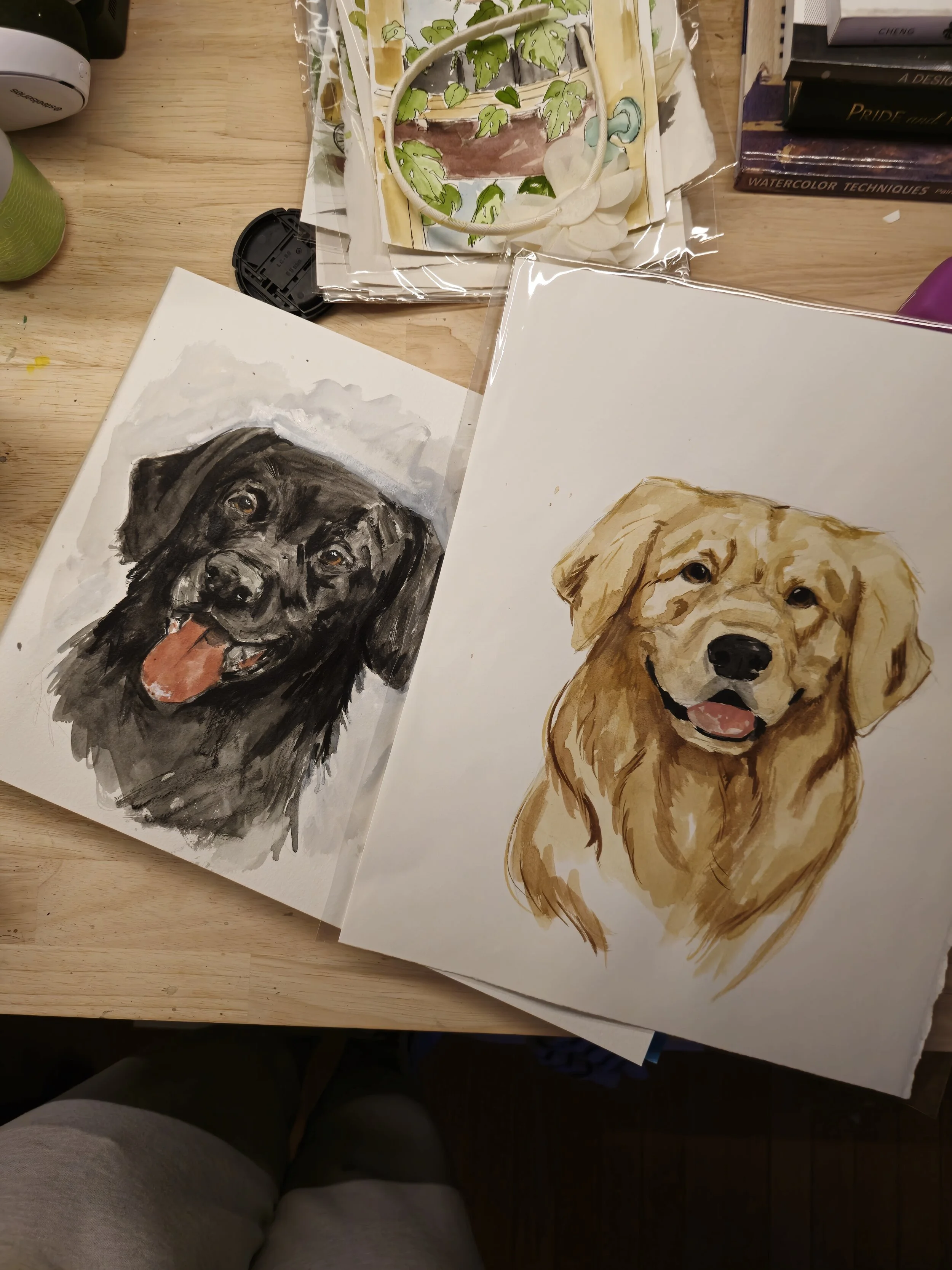 Two watercolor paintings of dogs on a wooden table, one black dog with an open mouth and tongue out, and one golden retriever smiling with its tongue out.