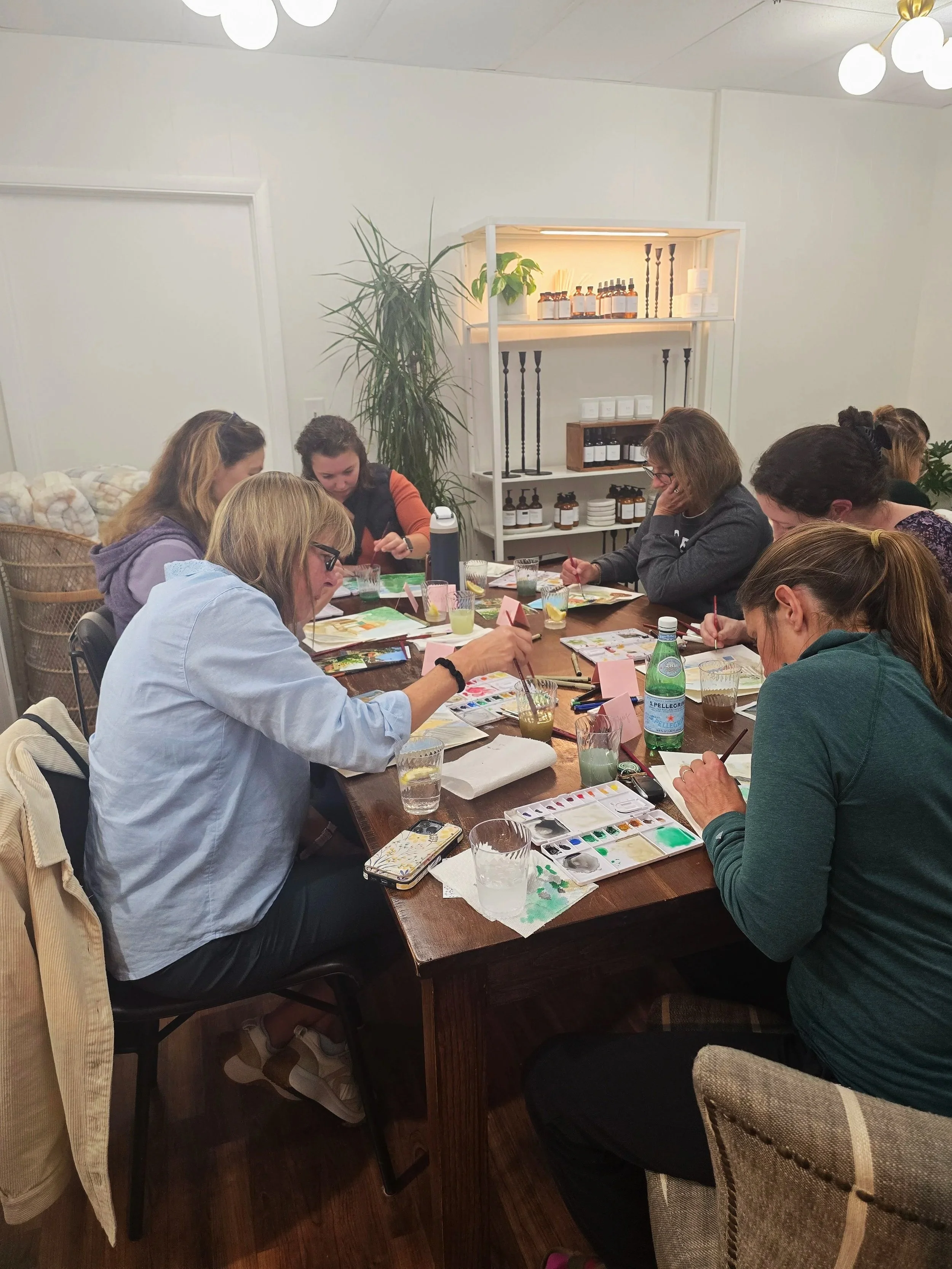 A group of women participating in a watercolor painting workshop around a wooden table. They have watercolor paints, brushes, and glasses of water. The room has white walls, a white shelf with bottles and candles, and room decor including a large plant.