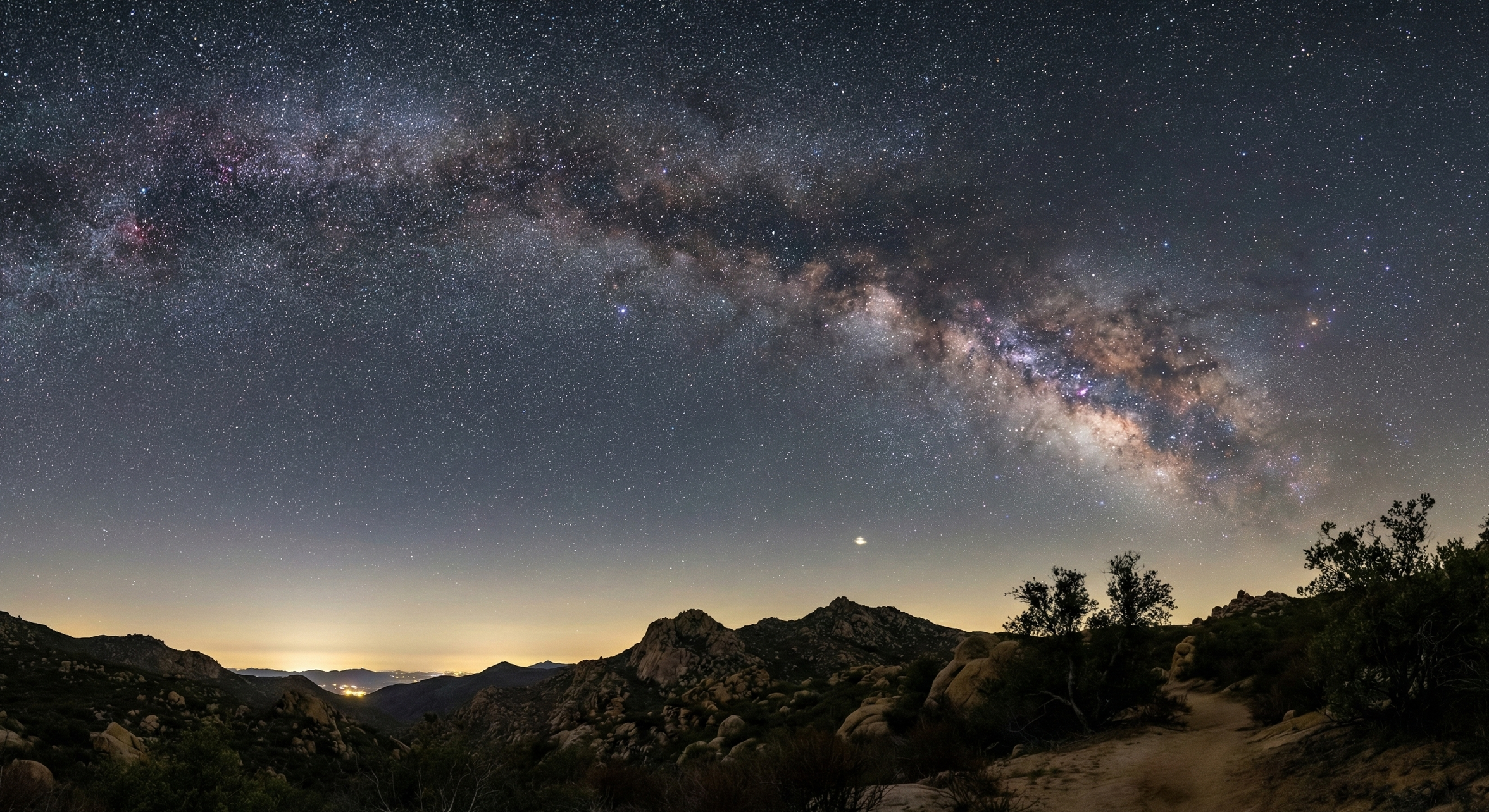 Night sky filled with stars and the Milky Way galaxy over a rugged desert landscape with rocks, bushes, and mountains.
