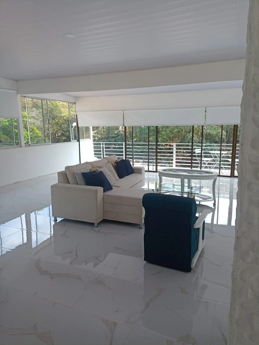 Bright living room with white marble floors, large windows, a white sofa with navy and cream pillows, a round glass-top table, and a blue armchair.