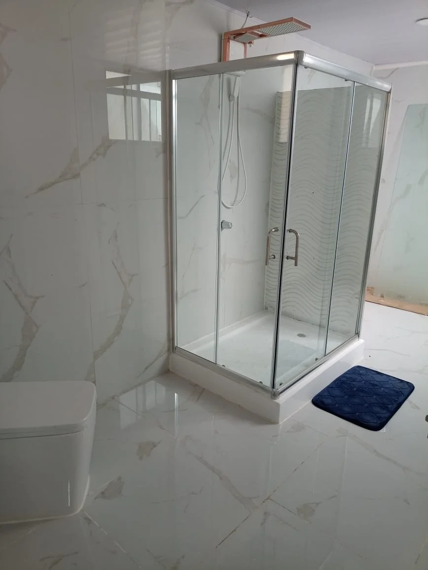 Bathroom with marble walls and floor, a glass shower enclosure with metal handles, a blue bath mat, and a white wall-mounted toilet.