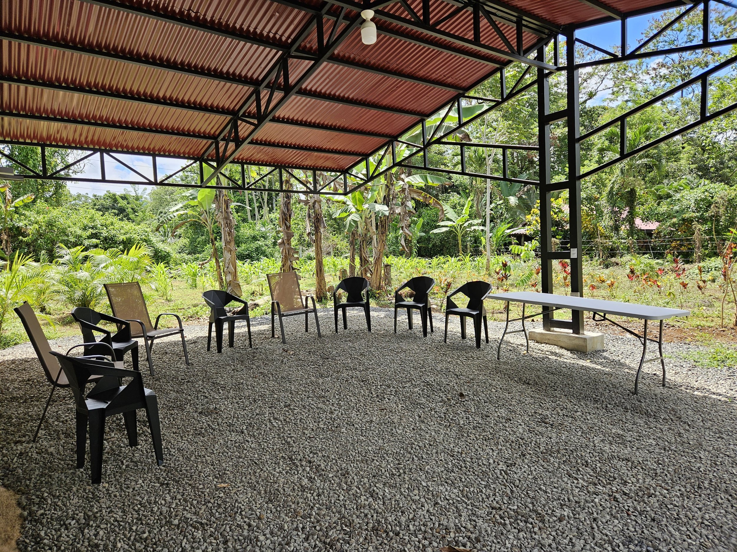 An outdoor covered seating area with eight chairs arranged in a semi-circle on a gravel ground, surrounded by lush green trees and plants, with a white folding table on the right side.