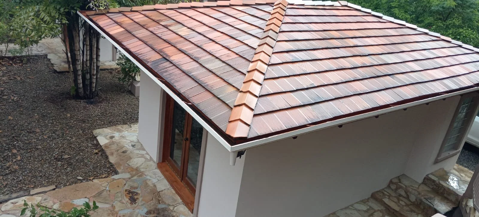 A roof with reddish-brown tiles and a ridge cap, seen from above, attached to a house with sliding glass doors and stone steps outside.