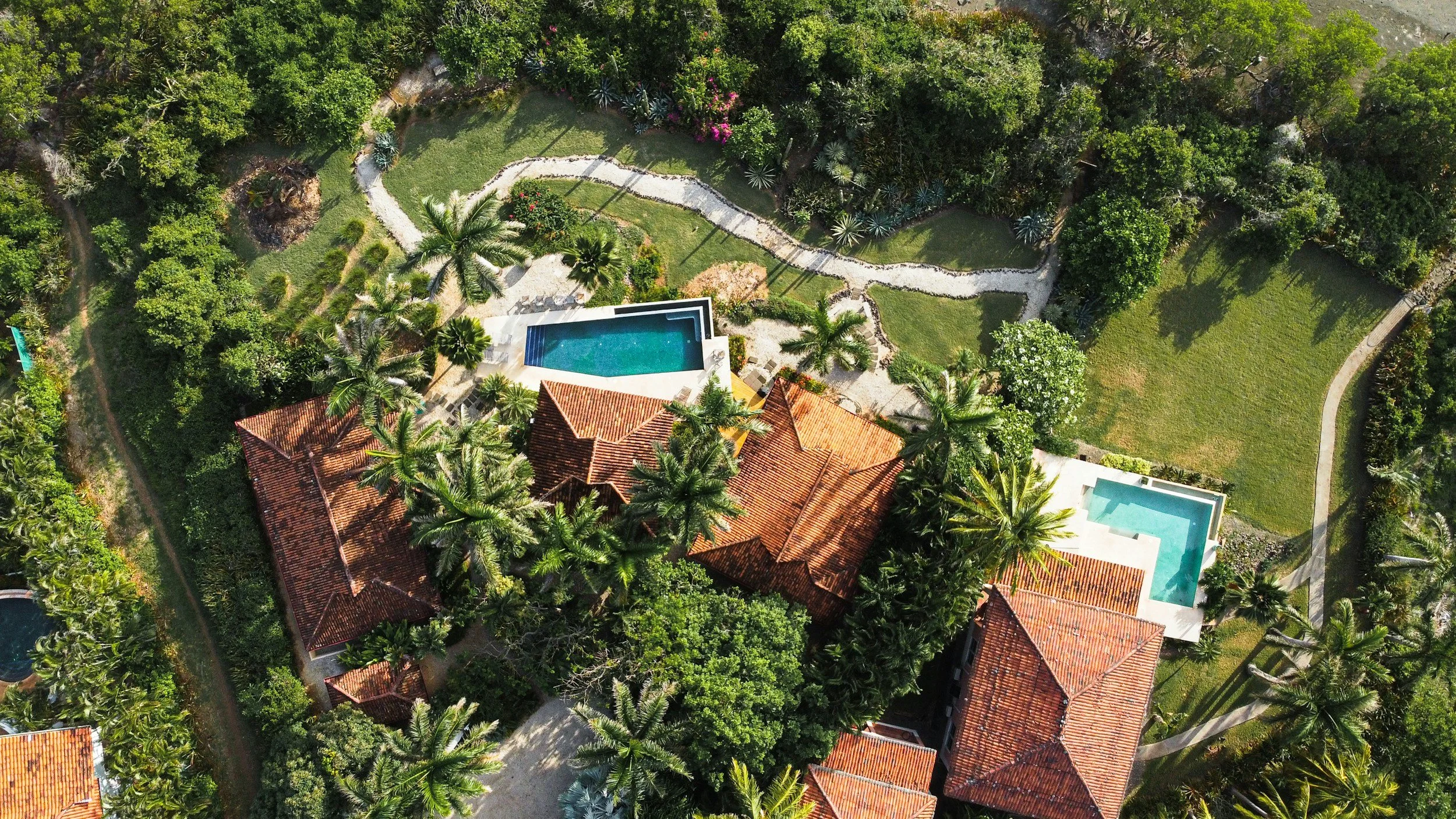 Aerial view of a lush green backyard with two swimming pools, surrounded by palm trees, pathways, and a house with a red tile roof.