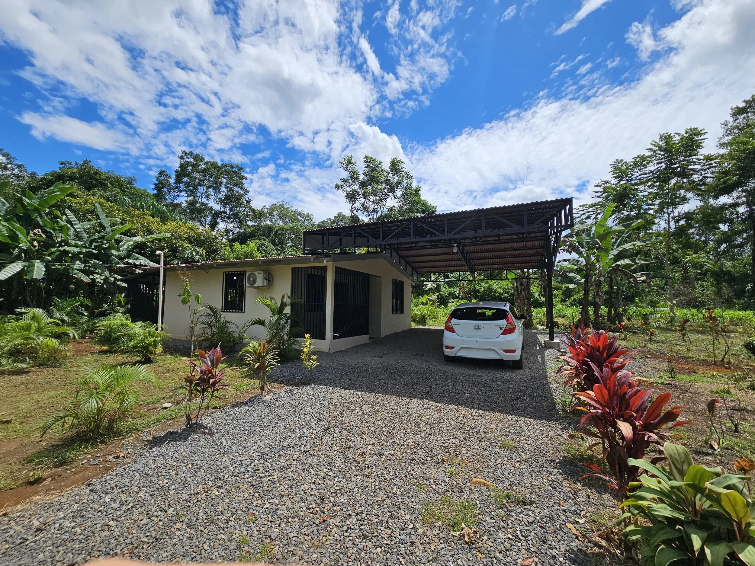 A small house with a carport, a white car parked underneath, and surrounded by lush green plants and trees, under a partly cloudy sky.