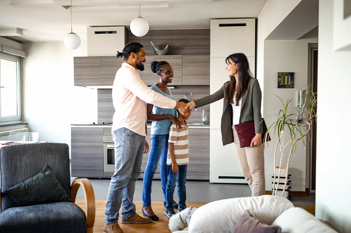 A family of four shaking hands with a woman in an apartment kitchen, with a dog sitting nearby.