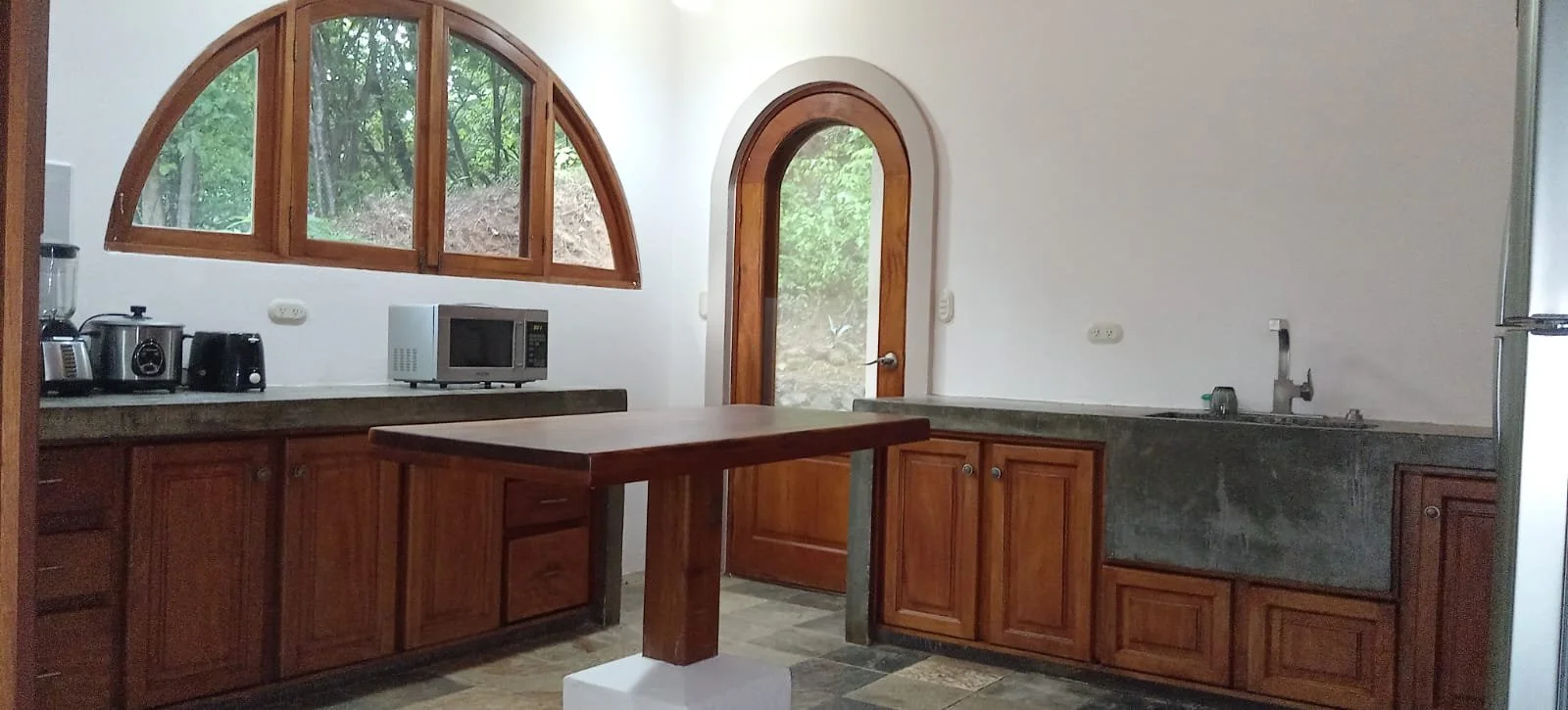 Kitchen with wooden cabinets, a microwave, blender, and coffee maker on the counter, arched and rectangular windows with green trees outside, door with glass panel, granite sink with faucet, and a small kitchen island.