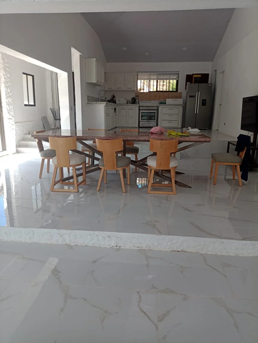 Open-concept kitchen and dining area with a marble floor, white cabinets, a stainless steel refrigerator, a large wooden dining table, and chairs. A window with blinds is visible above the sink.
