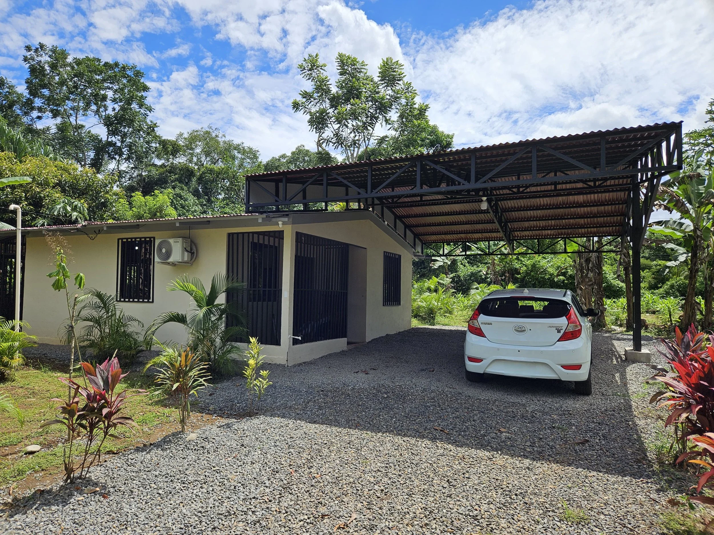 A small house with a carport, a white hatchback, and surrounding greenery under a partly cloudy sky.