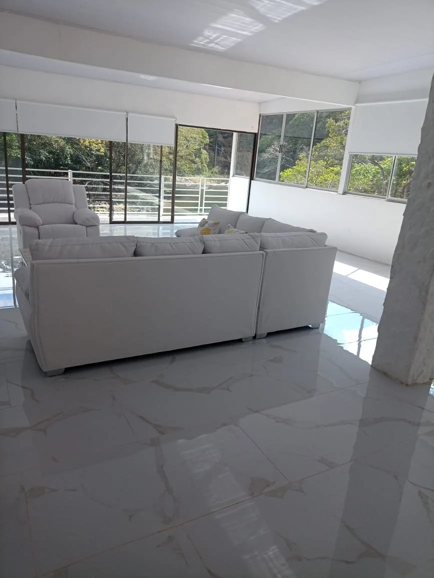 Bright living room with white couches and armchair, large windows showing trees outside, marble floor, and white blinds.