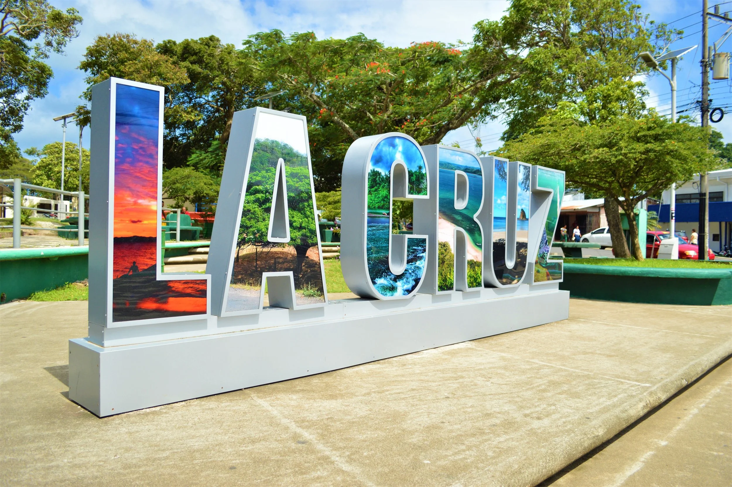 Large white 3D letters spelling 'LA CRUZ' with scenic tropical beach and ocean photos inside the letters, set in a park with trees, benches, and people in the background.