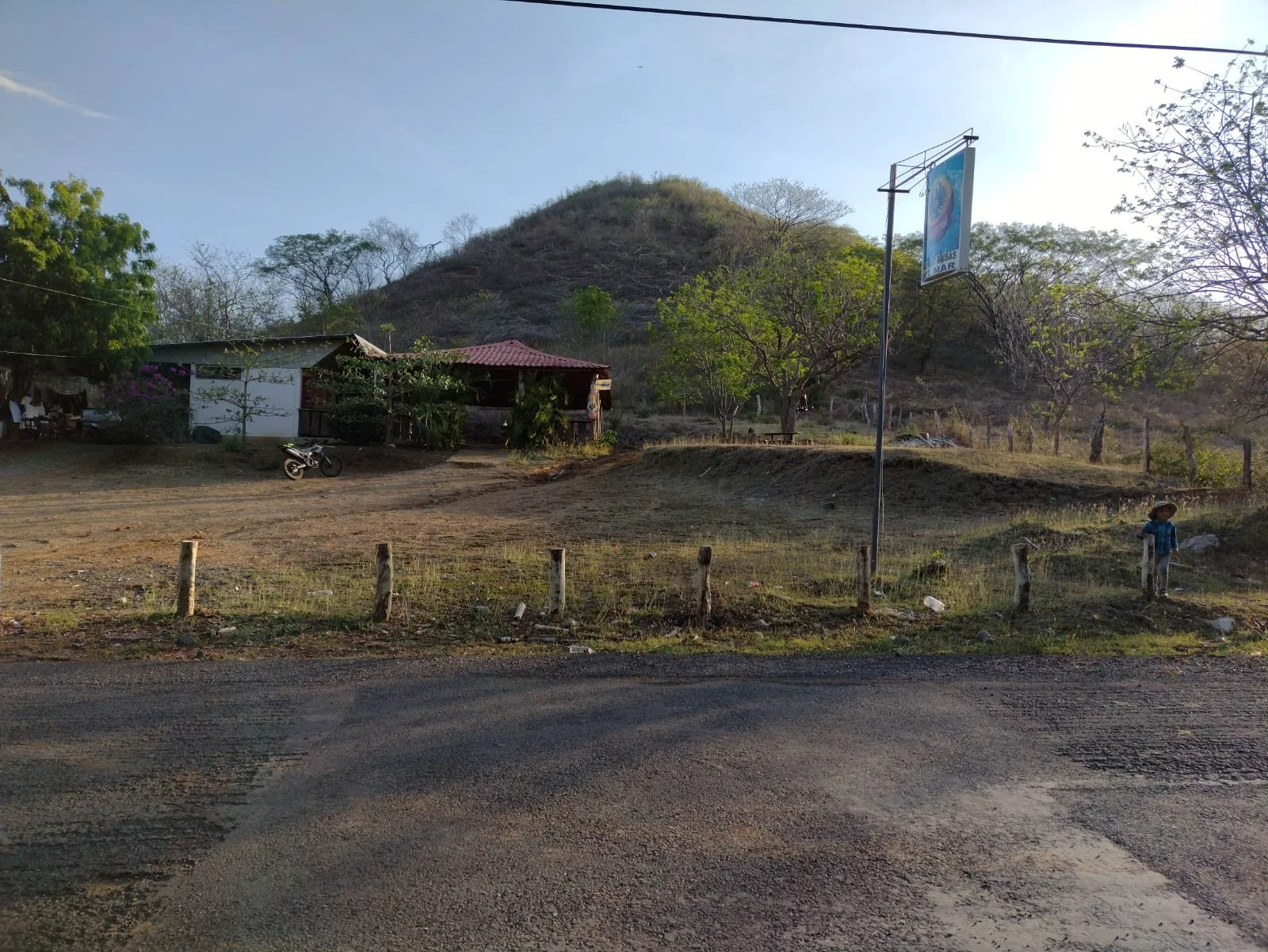 A rural scene with a dirt road, a small house, a motorcycle, a person with a hat on the right, a hill in the background, and a sky with some clouds.