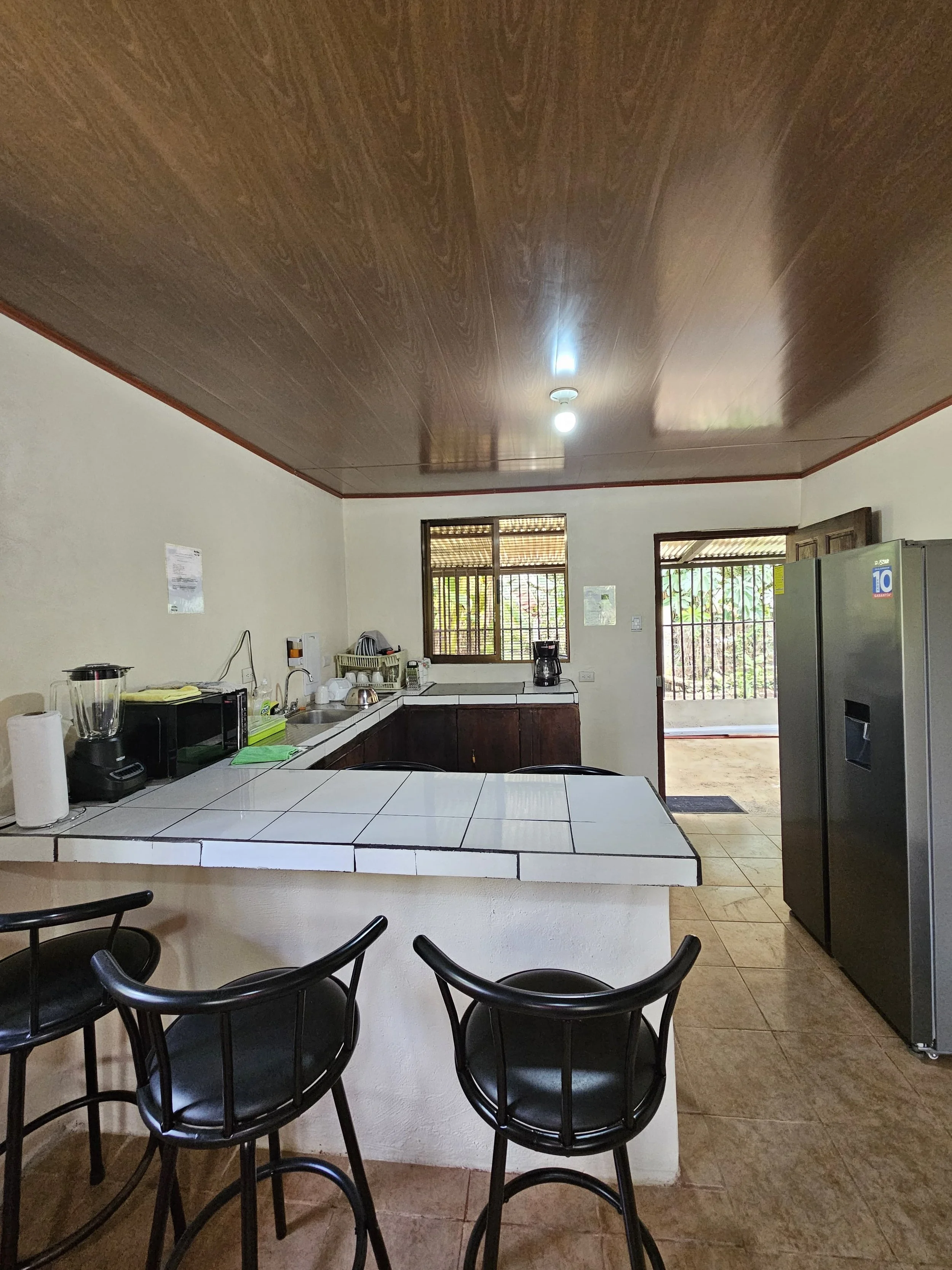 Kitchen with tiled countertop, bar stools, microwave, blender, coffee maker, refrigerator, cabinets, windows, and door.
