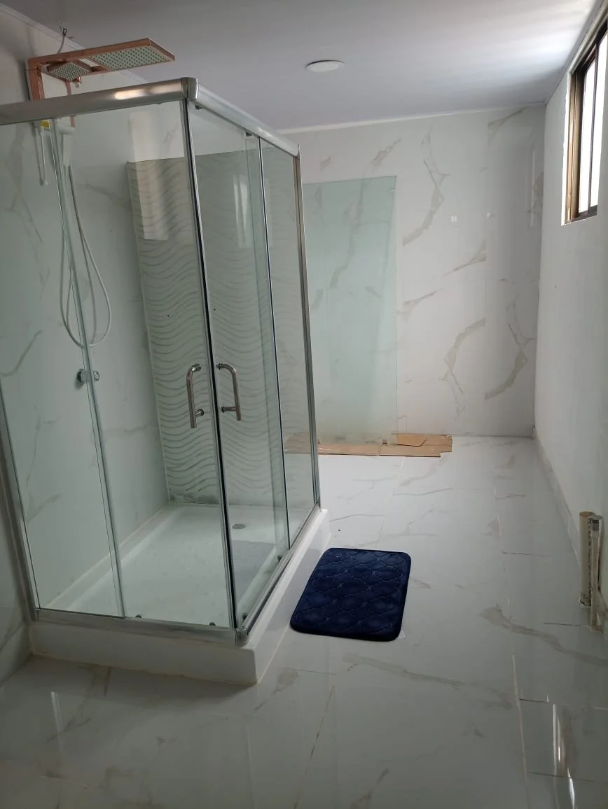Empty bathroom with a glass shower enclosure, a small black bath mat, and a window on the right wall. The bathroom appears to be under renovation.