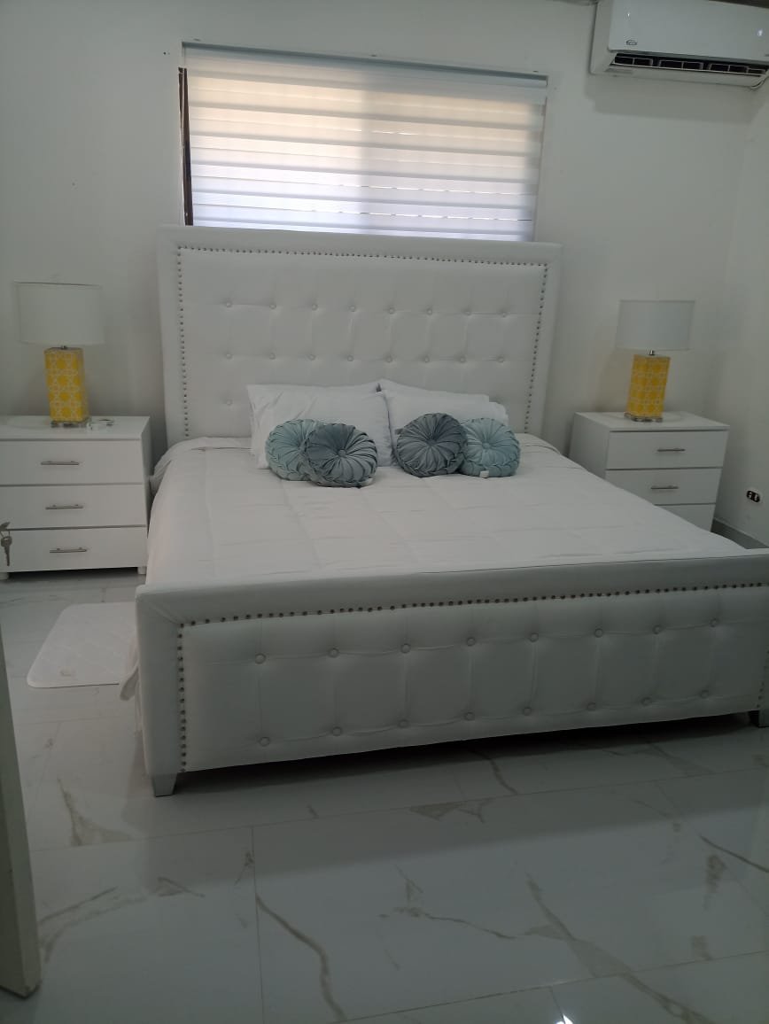 A bedroom with a white upholstered bed, white nightstands with lamps, and decorative gray pillows, against light-colored walls and a window with a white blind.