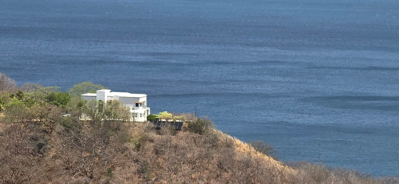 A coastal scene with a white house on a hillside covered with trees and shrubs, overlooking a large body of water.