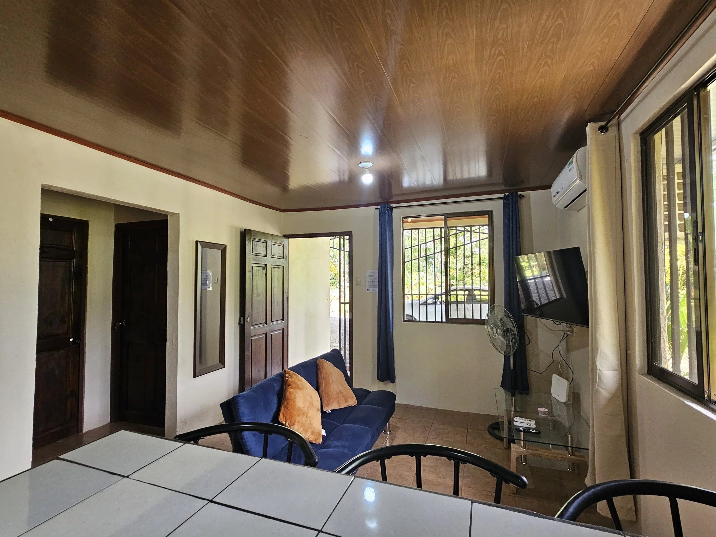 Living room with wooden ceiling, beige walls, a glass-top table, a navy blue couch with tan cushions, window with blue curtains, a wall-mounted flat-screen TV, a standing fan, and an air conditioner.