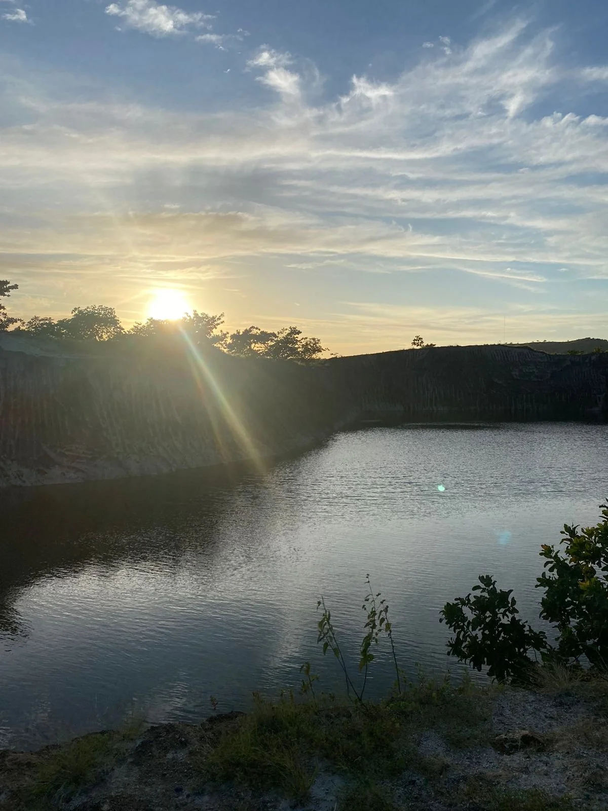 Sunset over a small lake with trees and hills in the background, partly cloudy sky.