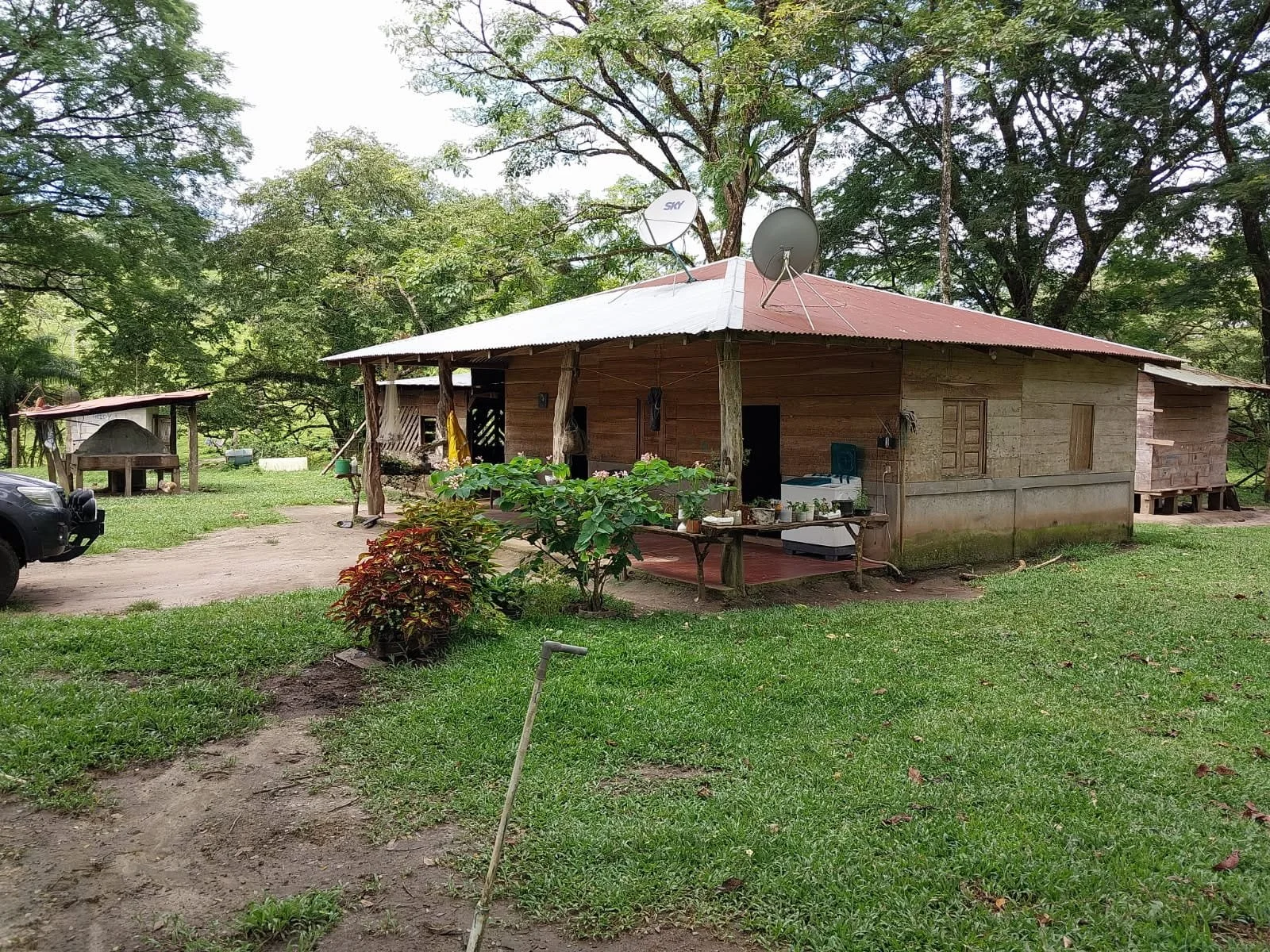 A rustic wooden house with a tin roof, surrounded by trees and greenery. There are satellite dishes on the roof, a small porch with plants, and a driveway with a black vehicle parked nearby.