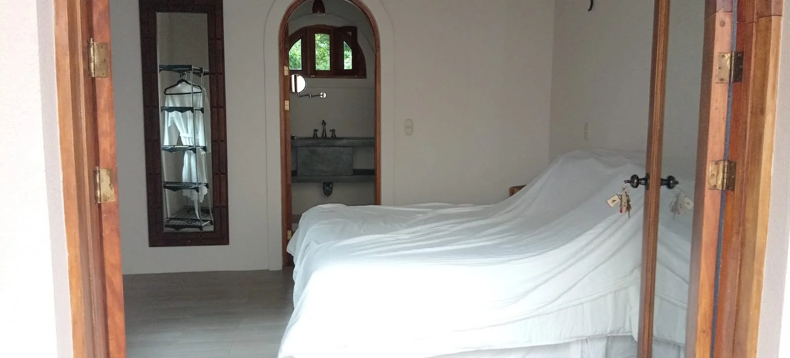 Bedroom with a bed covered in a white sheet, a mirror on the wall, a laundry rack, and a small sink with a window above it.