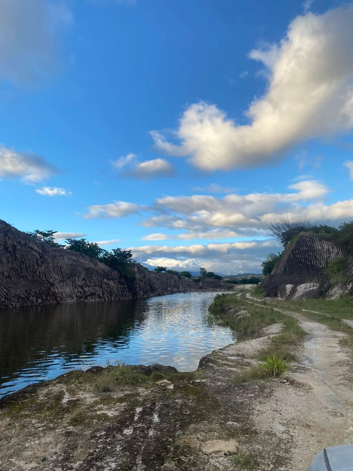 River flowing through canyon with rocky sides; dirt road parallel to river on right; blue sky with scattered clouds.