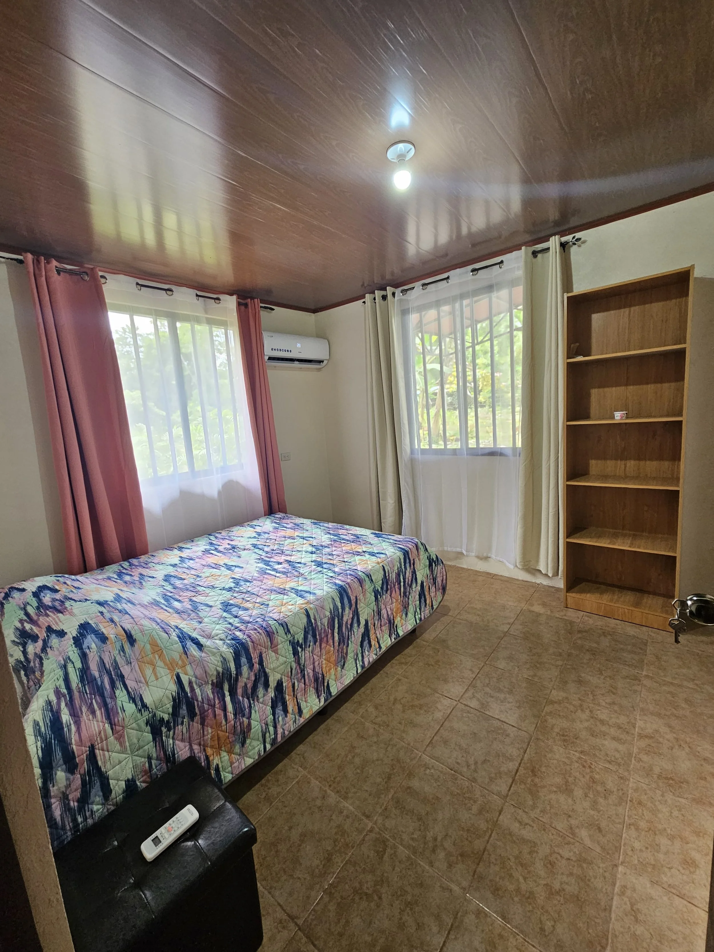 A small bedroom with a wooden ceiling, two windows with white curtains, a bed with colorful patterned bedding, a black storage ottoman with a remote on top, a wooden bookshelf, and an air conditioning unit on the wall.